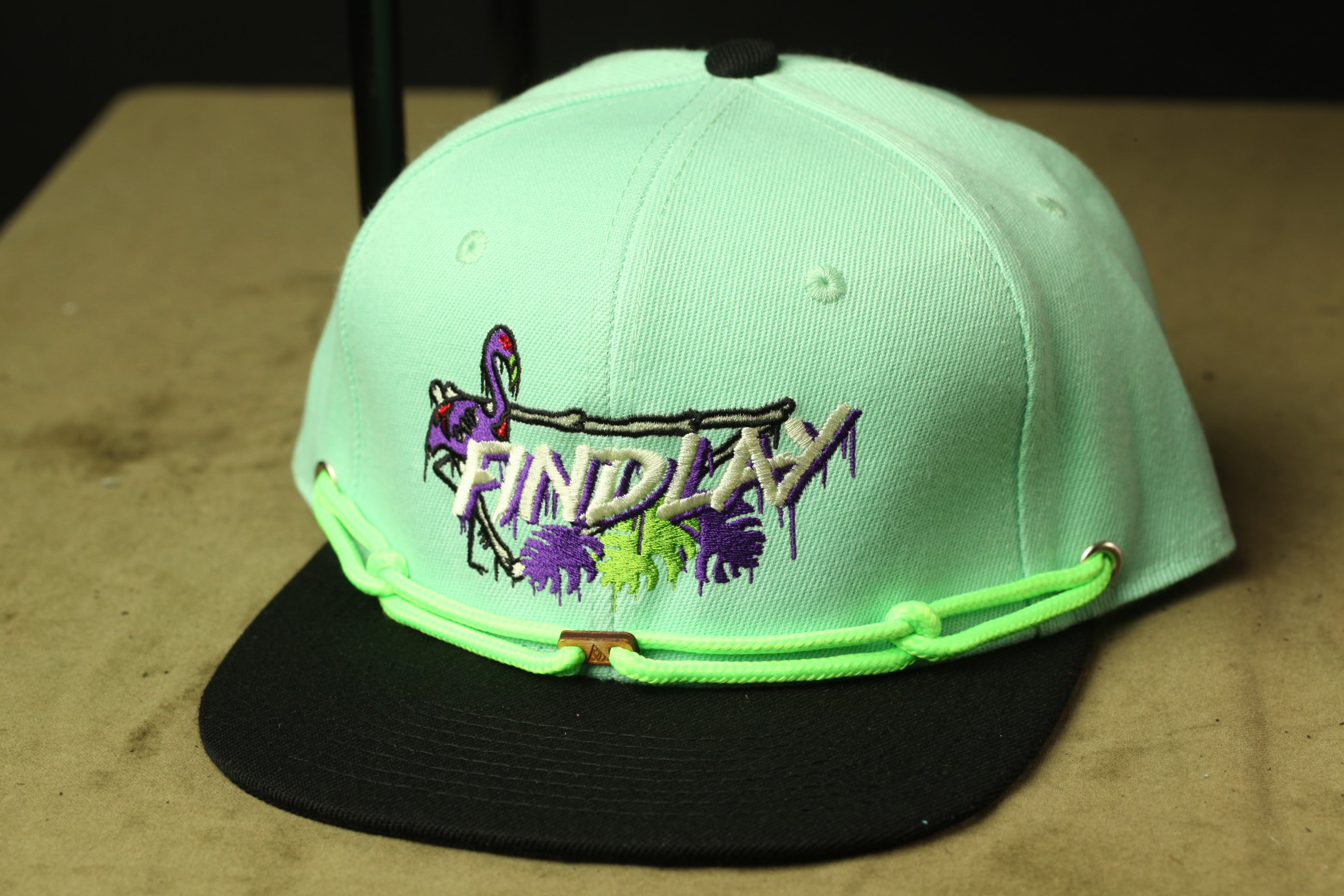 A mint green baseball cap with a black brim and a green string around the brim. The cap has a graphic design on the front, which includes a purple flamingo and the word "FINDlay" written in purple letters.