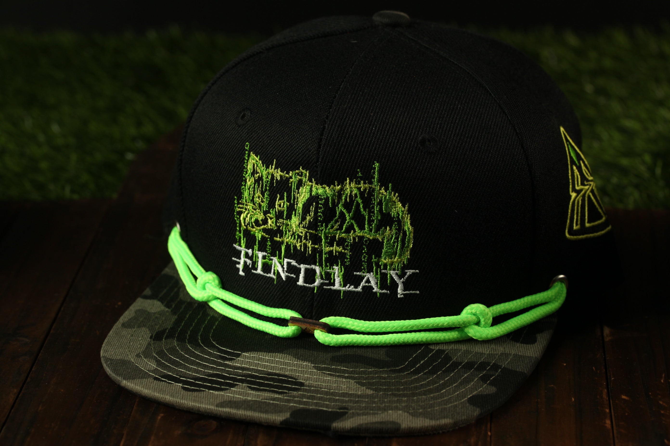 A black baseball cap with a camouflage pattern and a green string around the brim.