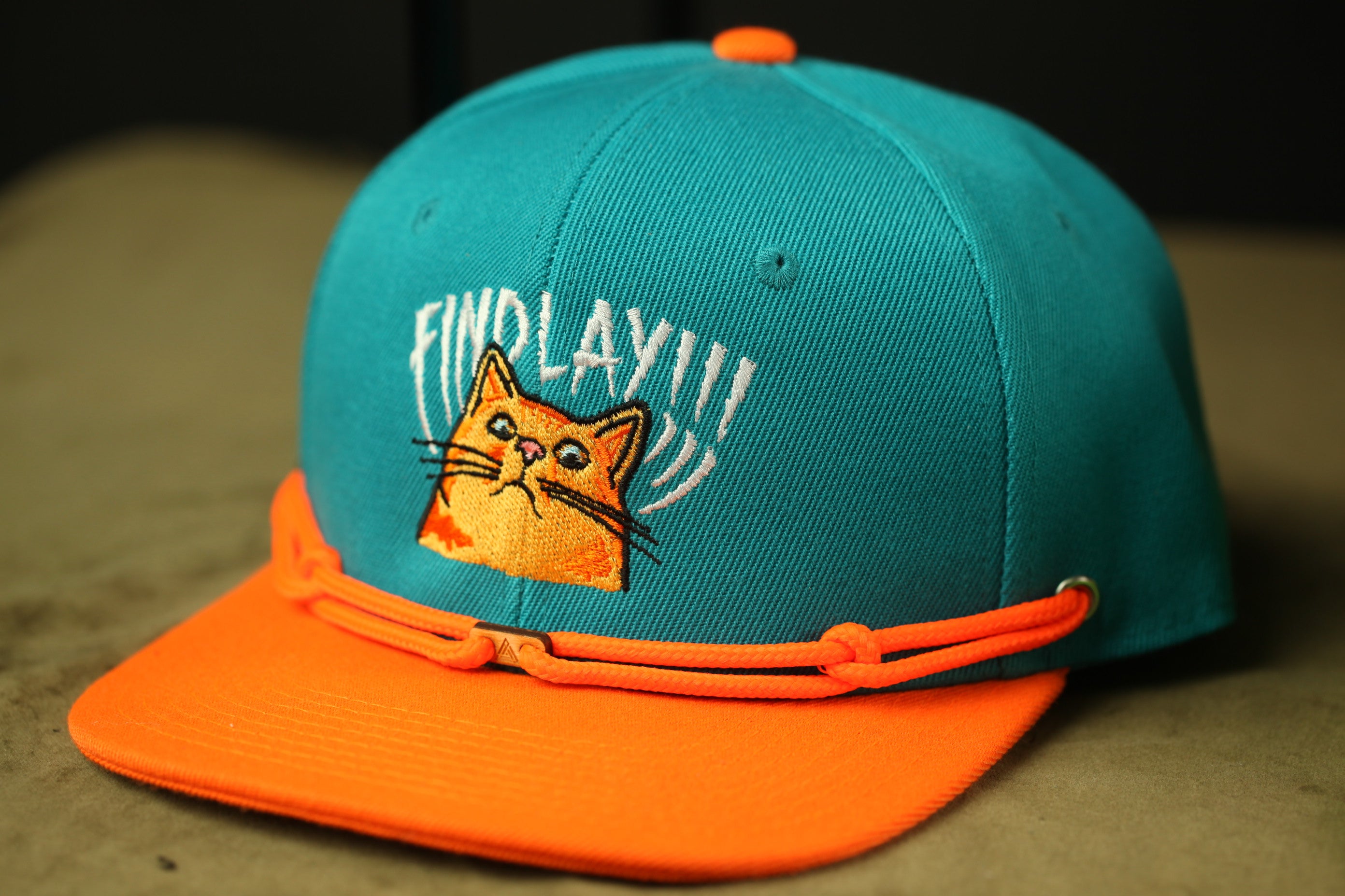 A turquoise baseball cap with an orange brim and a cat embroidered on the front.