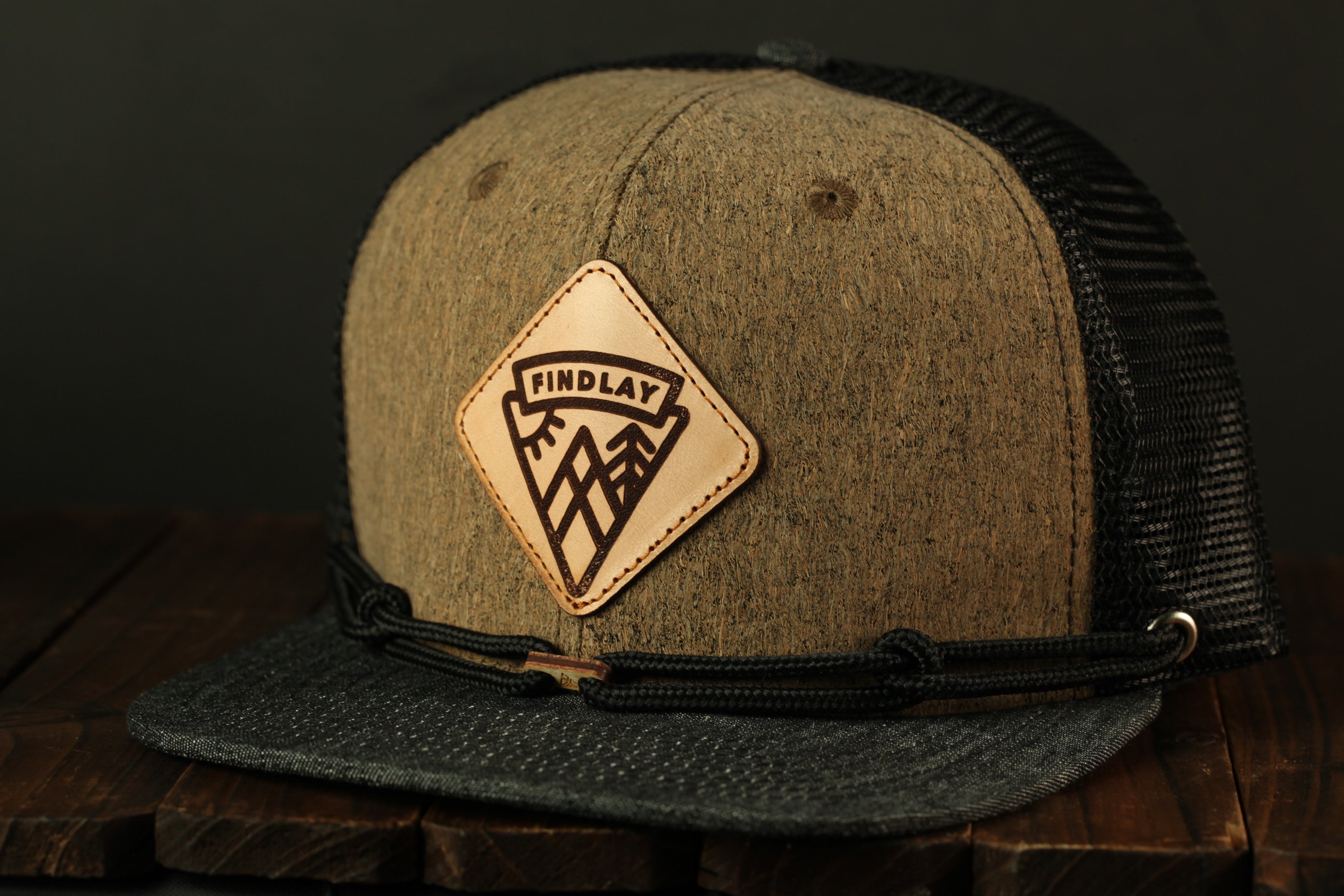 A gray and black baseball cap with a leather patch on the front, featuring a mountain and tree design.