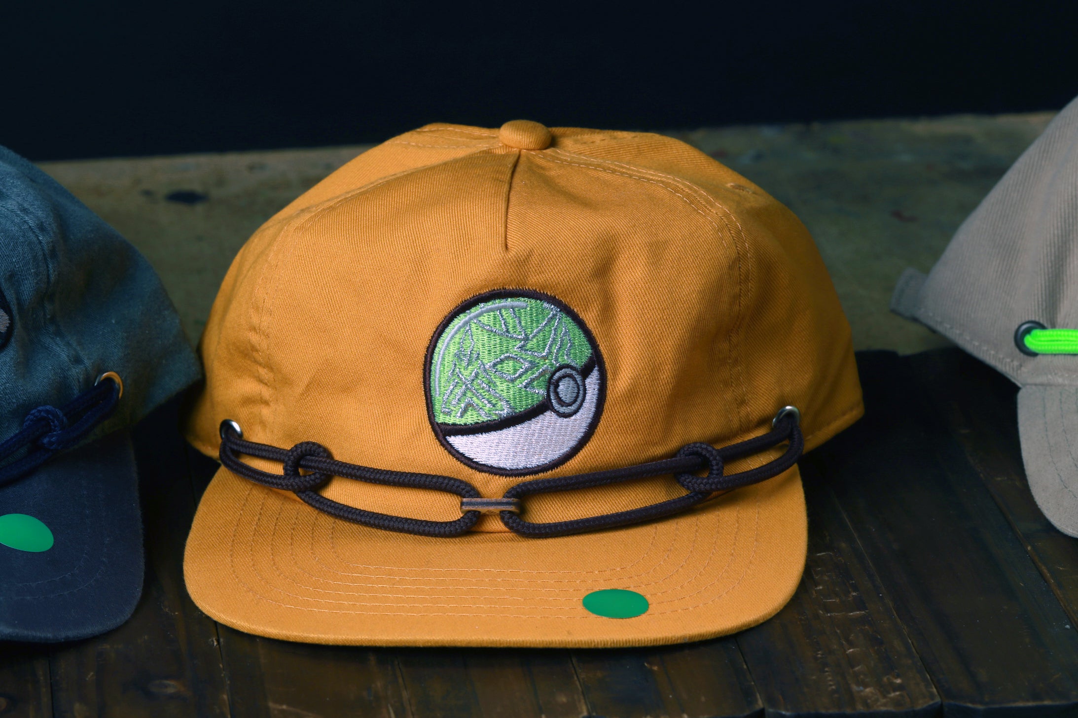 A yellow baseball cap with a green patch and a chain strap is displayed on a wooden surface, with other hats visible in the background.