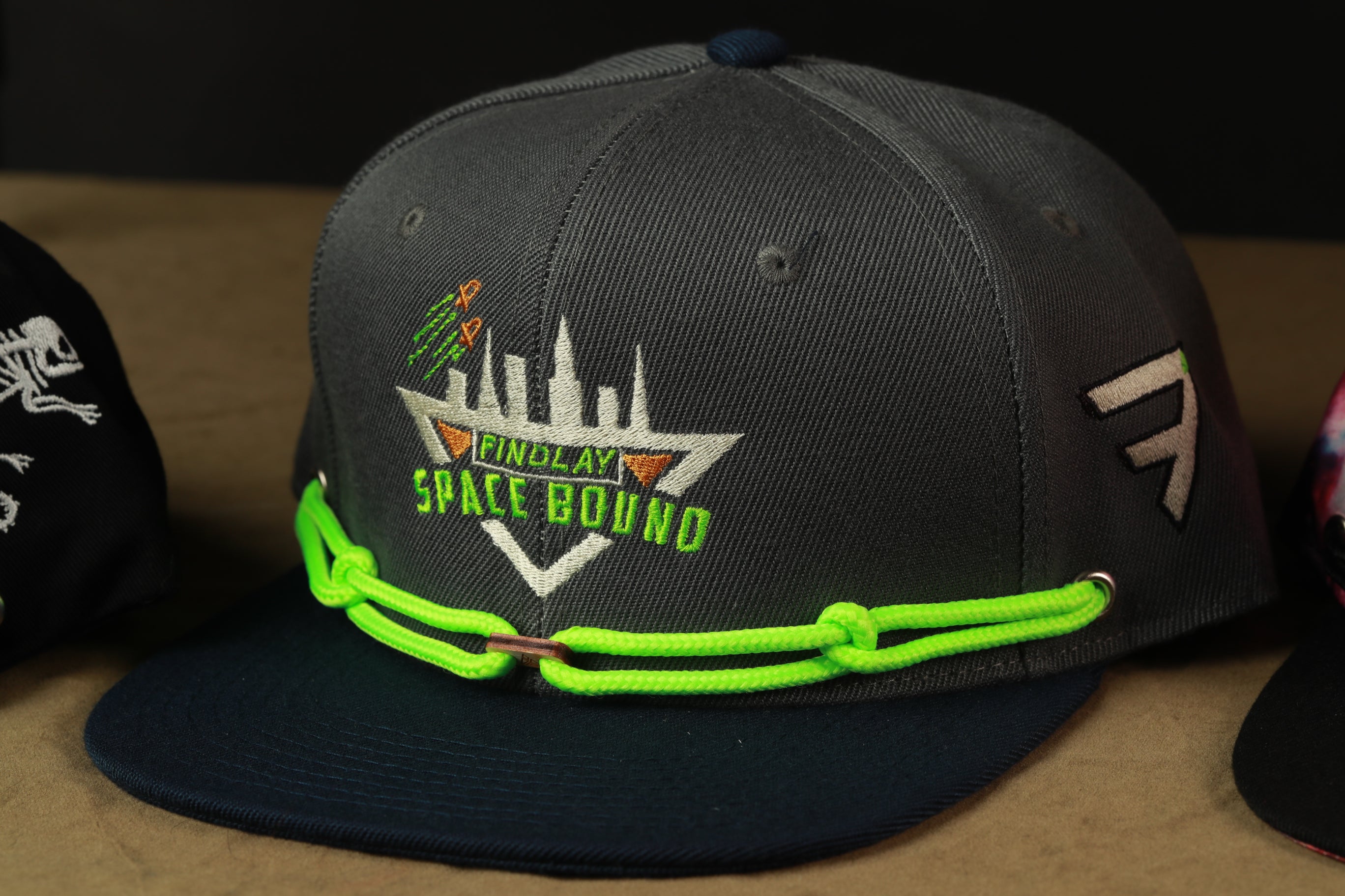 A close-up image of a gray baseball cap with a neon green chain strap, featuring a logo and text on the front.