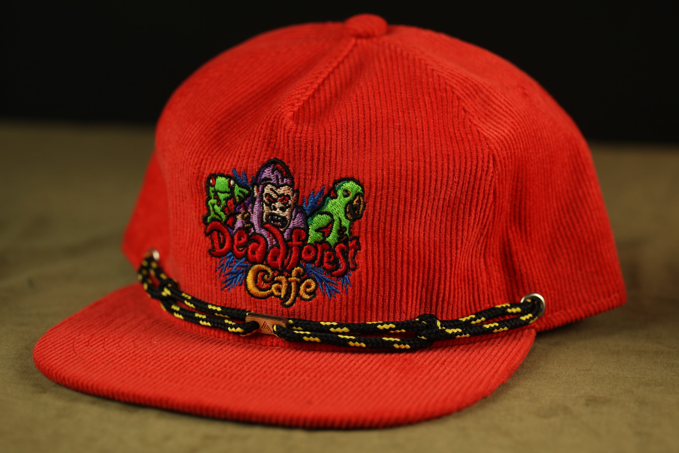 A red baseball cap with a black and yellow braided strap, featuring a colorful embroidered logo on the front.