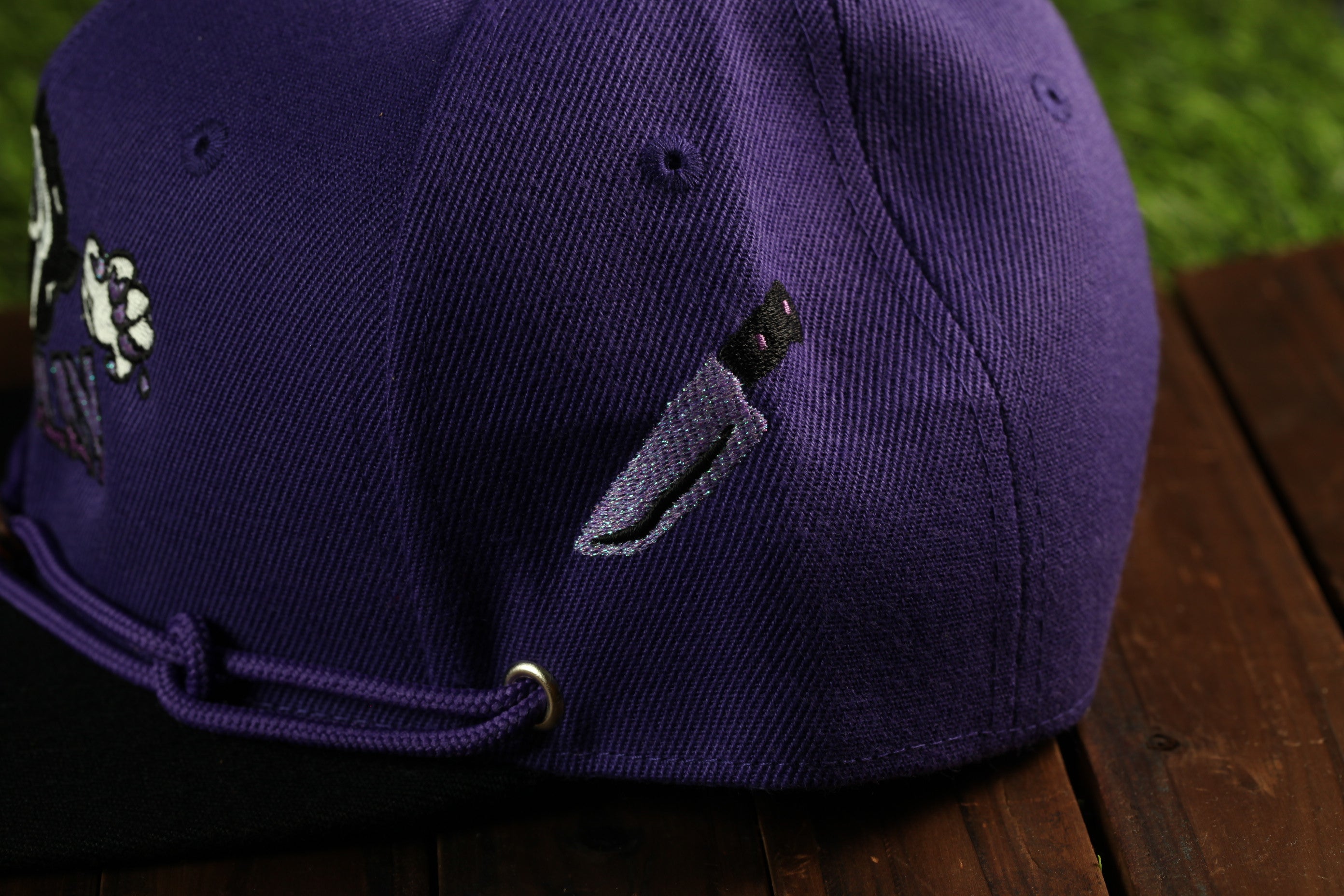 A close-up view of a purple baseball cap with a knife embroidered on it, resting on a wooden surface.