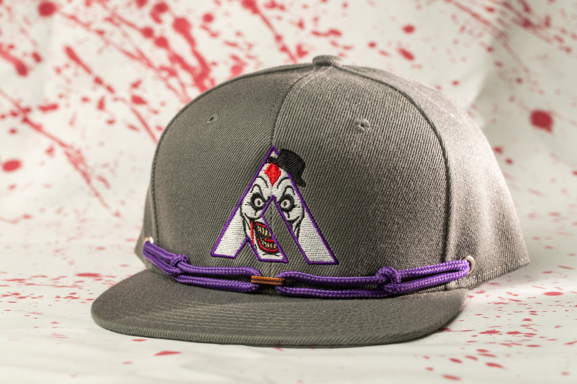A gray baseball cap with a purple string and a clown-like logo on the front, set against a background with red splatters.