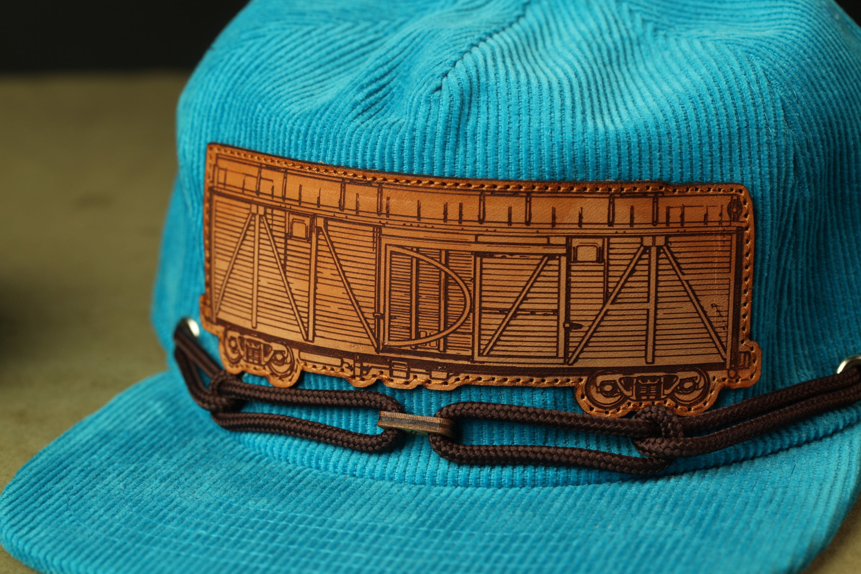 A close-up image of a blue baseball cap with a brown leather patch on the front, featuring an illustration of a train car.