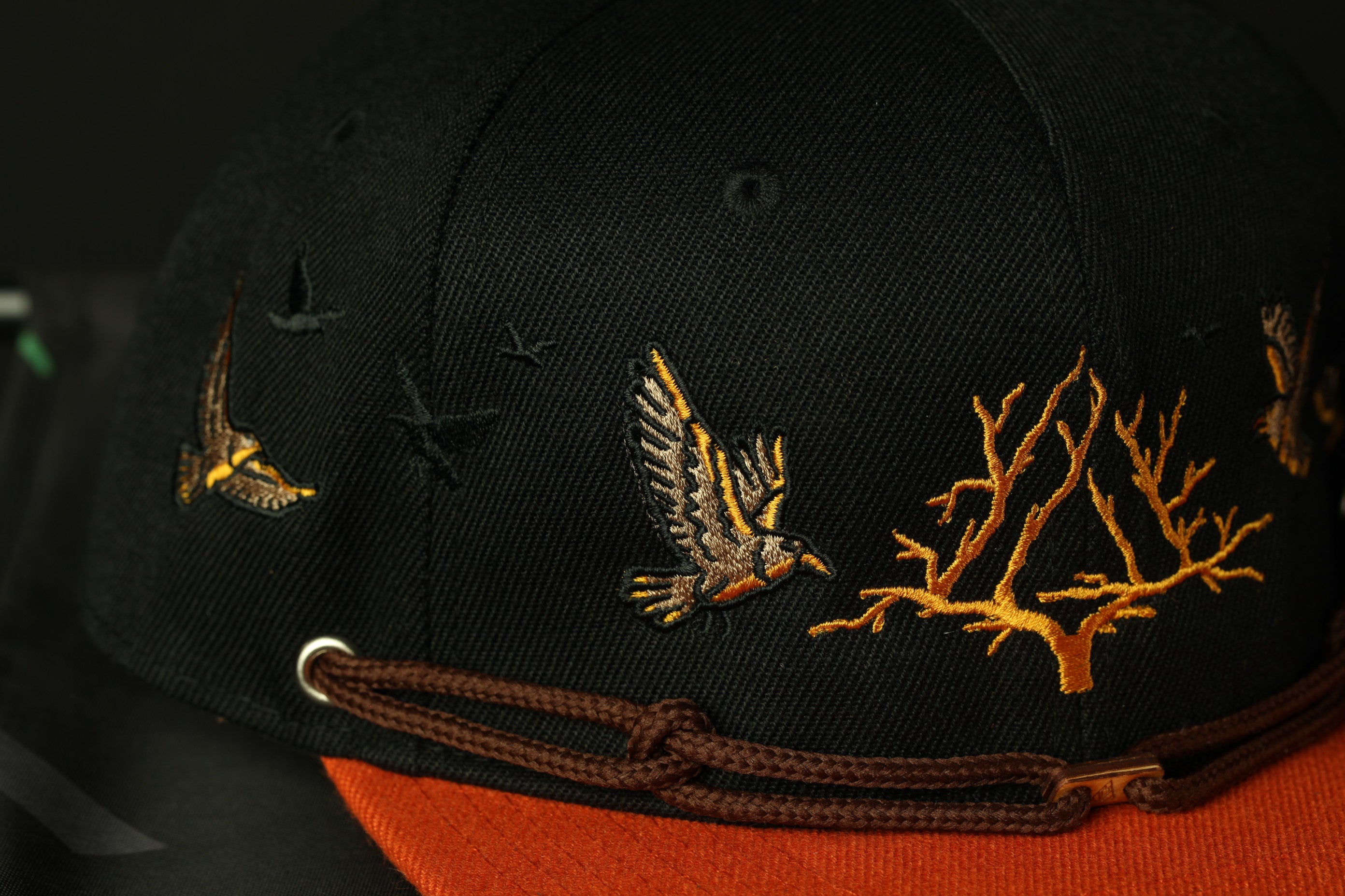 A close-up view of a black baseball cap with a brown strap and gold embroidered designs depicting birds and trees.