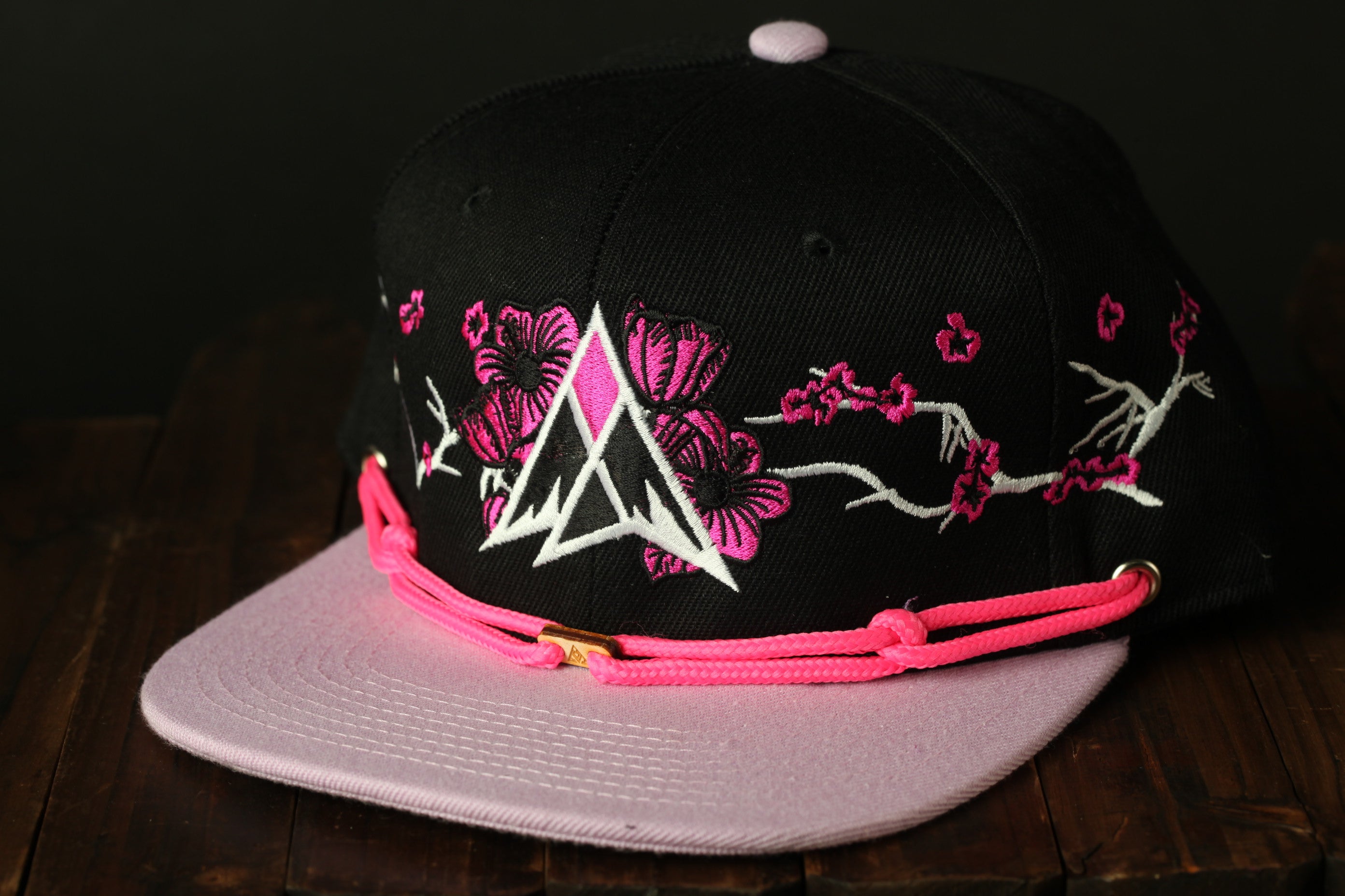 A black baseball cap with pink floral embroidery and a pink strap is displayed on a wooden surface.