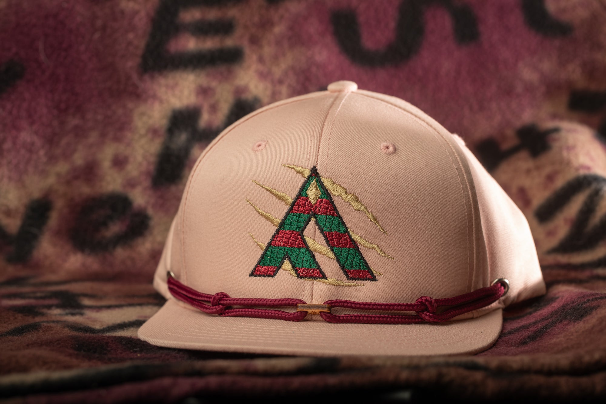 A pink baseball cap with a red and green embroidered logo on the front, placed on a patterned fabric background.