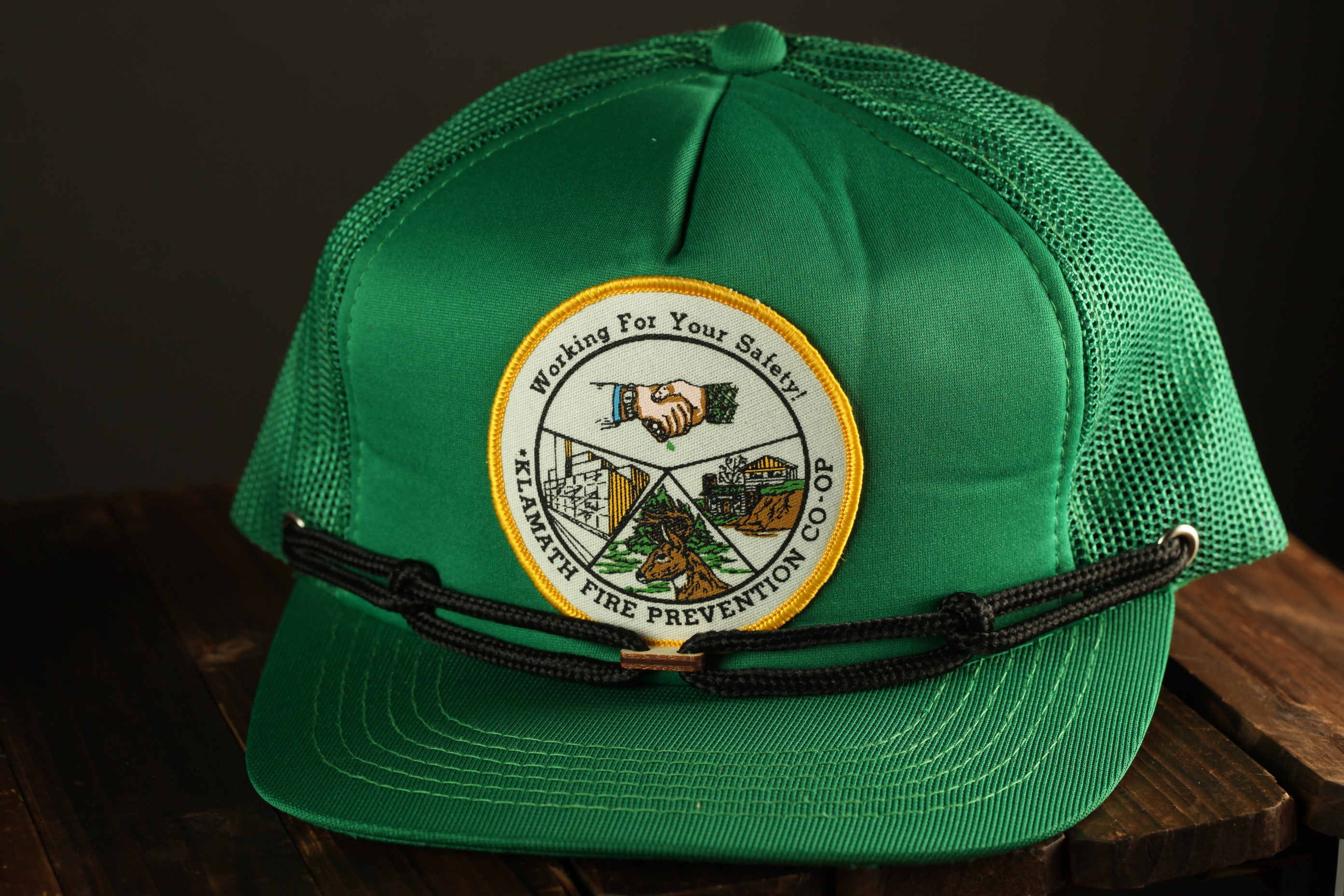 A green baseball cap with a patch on the front, featuring a handshake, a mountain, and text that says "Working For Your Safety! Klamath Fire Prevention Co.
