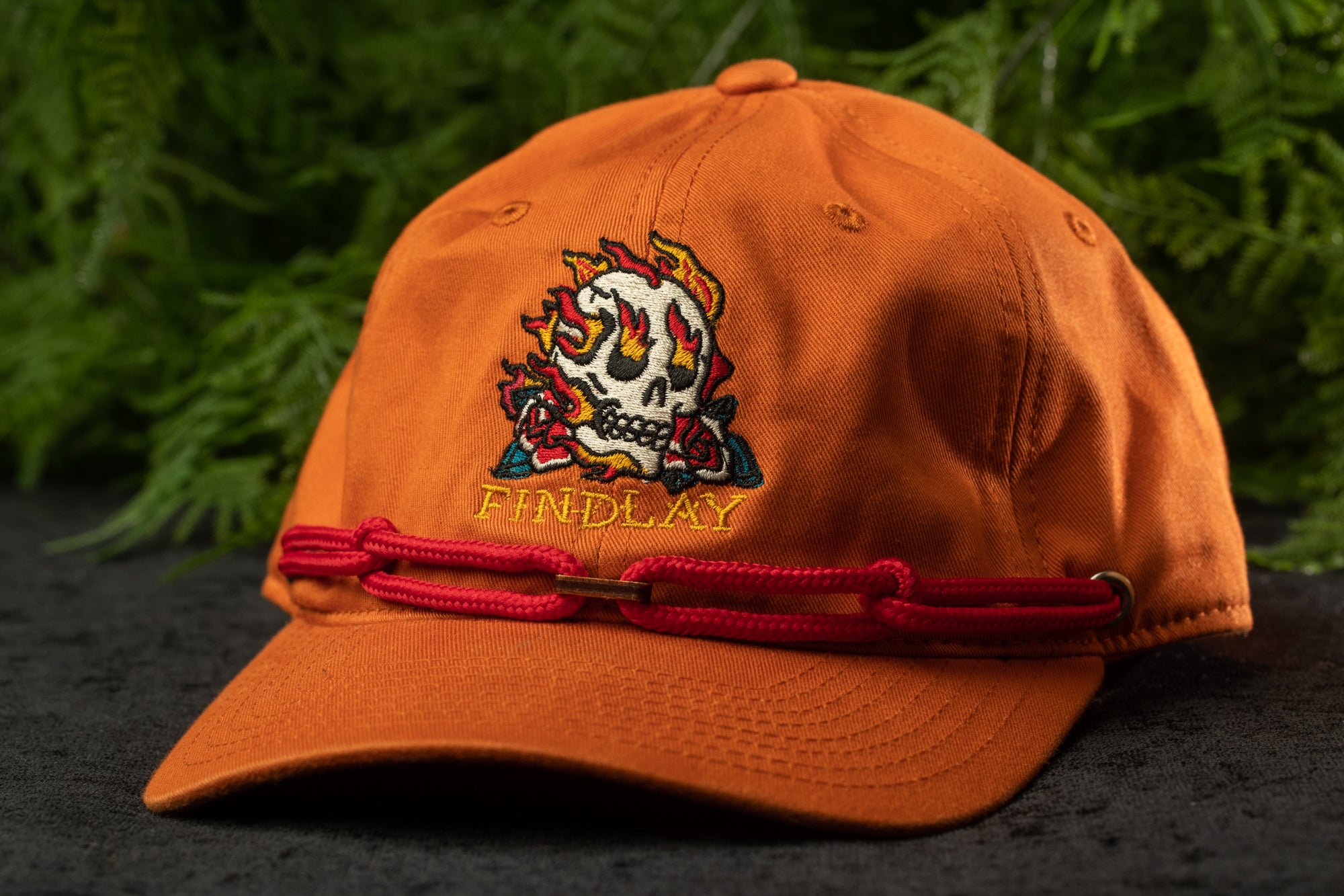 An orange baseball cap with a skull design and the word "FIND LAY" printed on it, placed on a dark surface with greenery in the background.