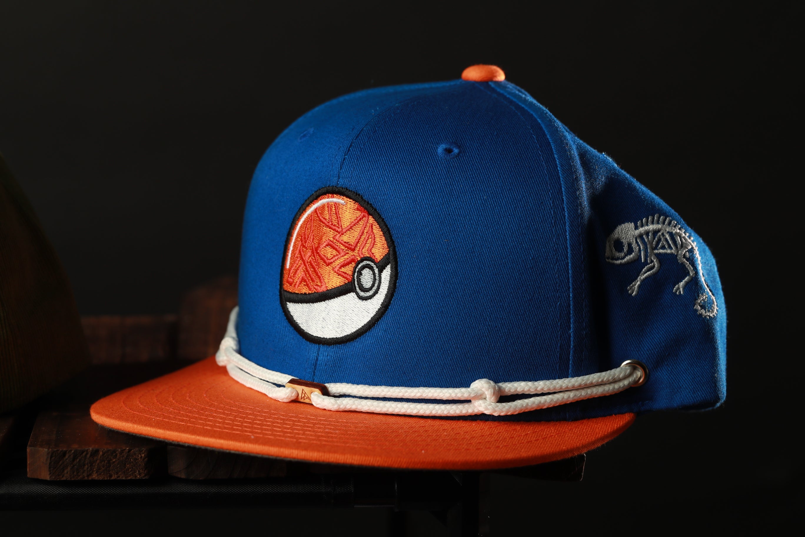 A blue baseball cap with an orange brim and a white Pokemon ball embroidered on the front, featuring a skeleton design on the right side.