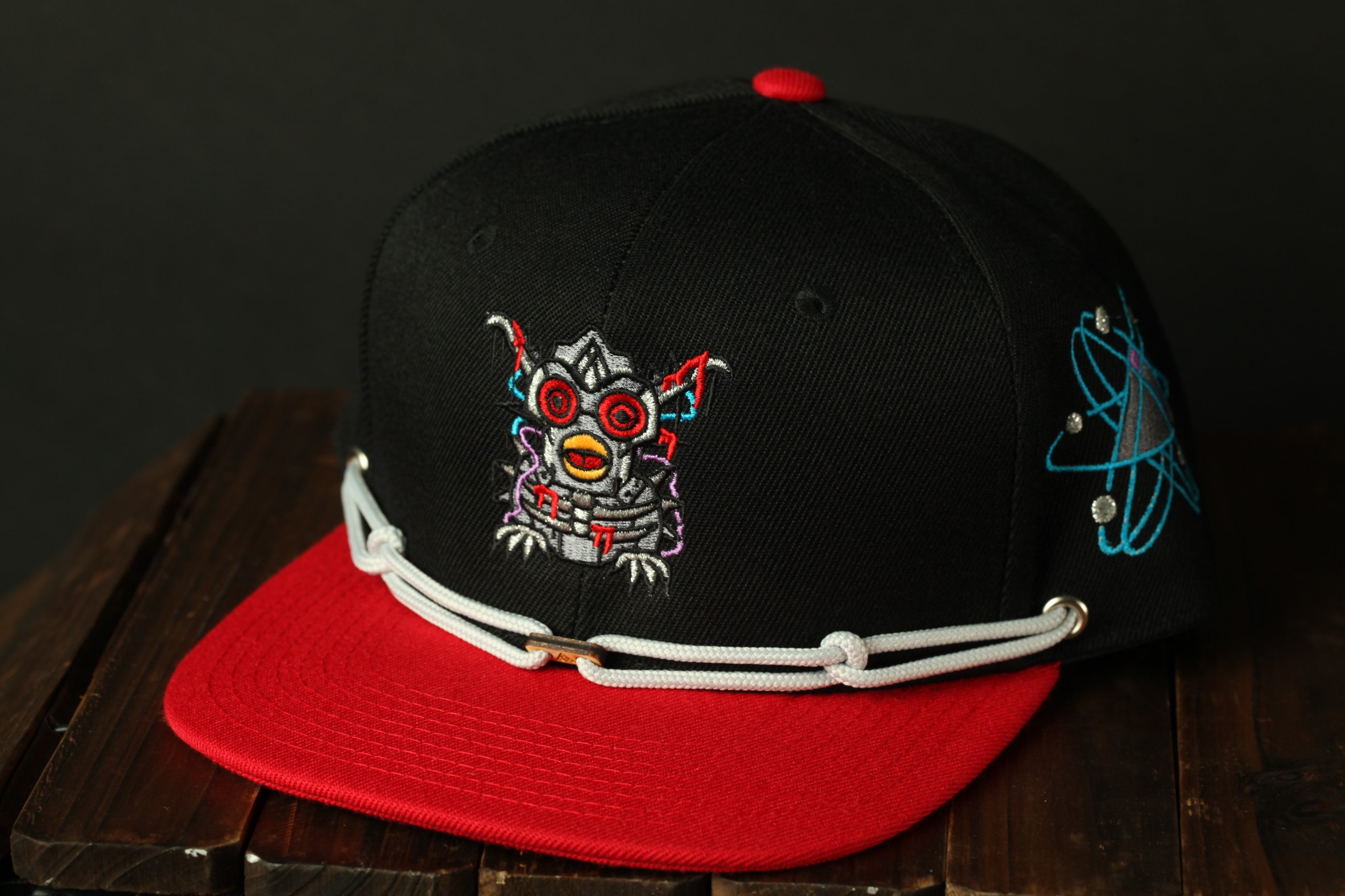 A black baseball cap with a red brim and a colorful, abstract design on the front, sitting on a wooden surface.