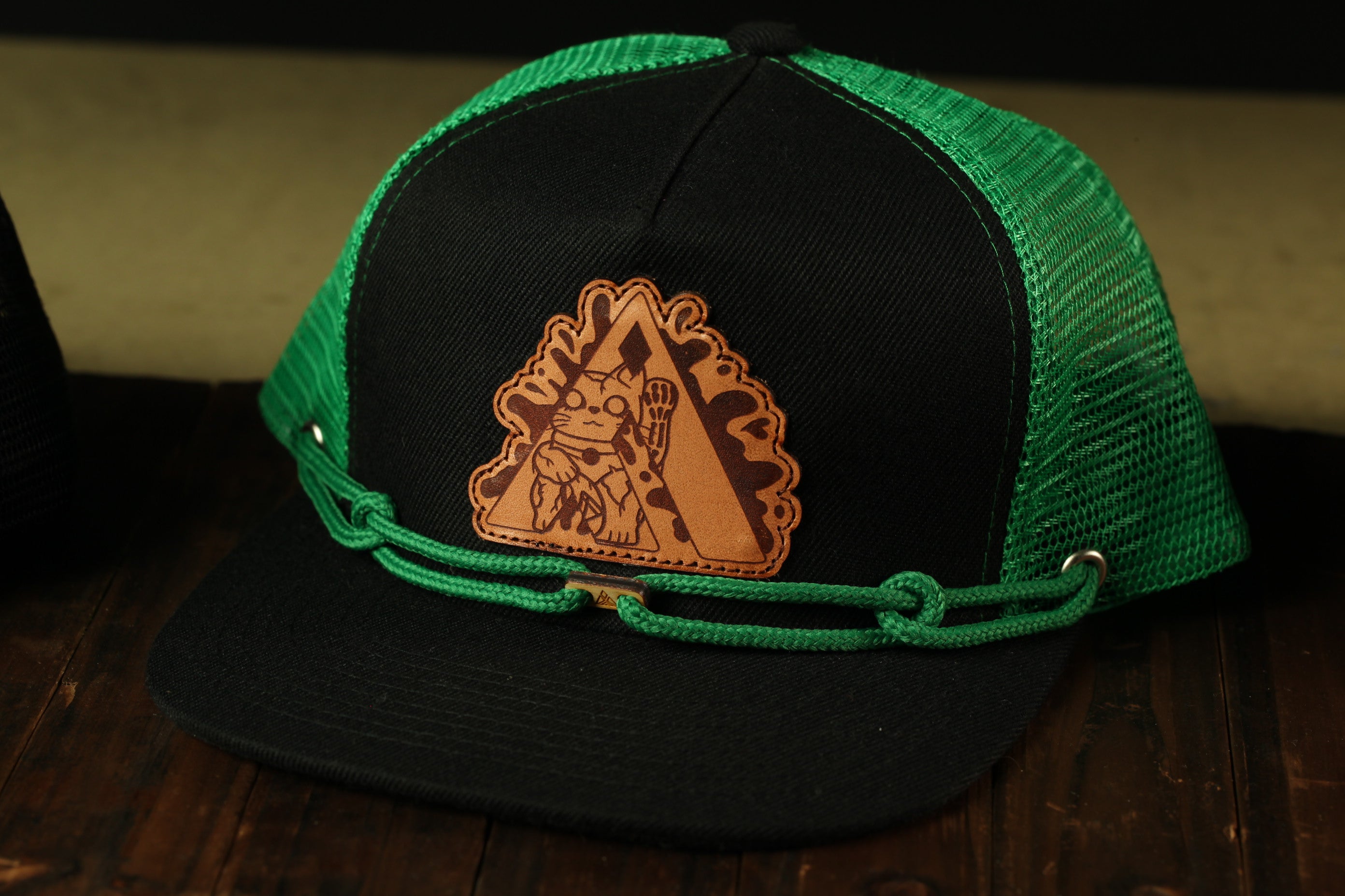 A black baseball cap with a green mesh panel and a brown leather patch featuring a cartoon character.
