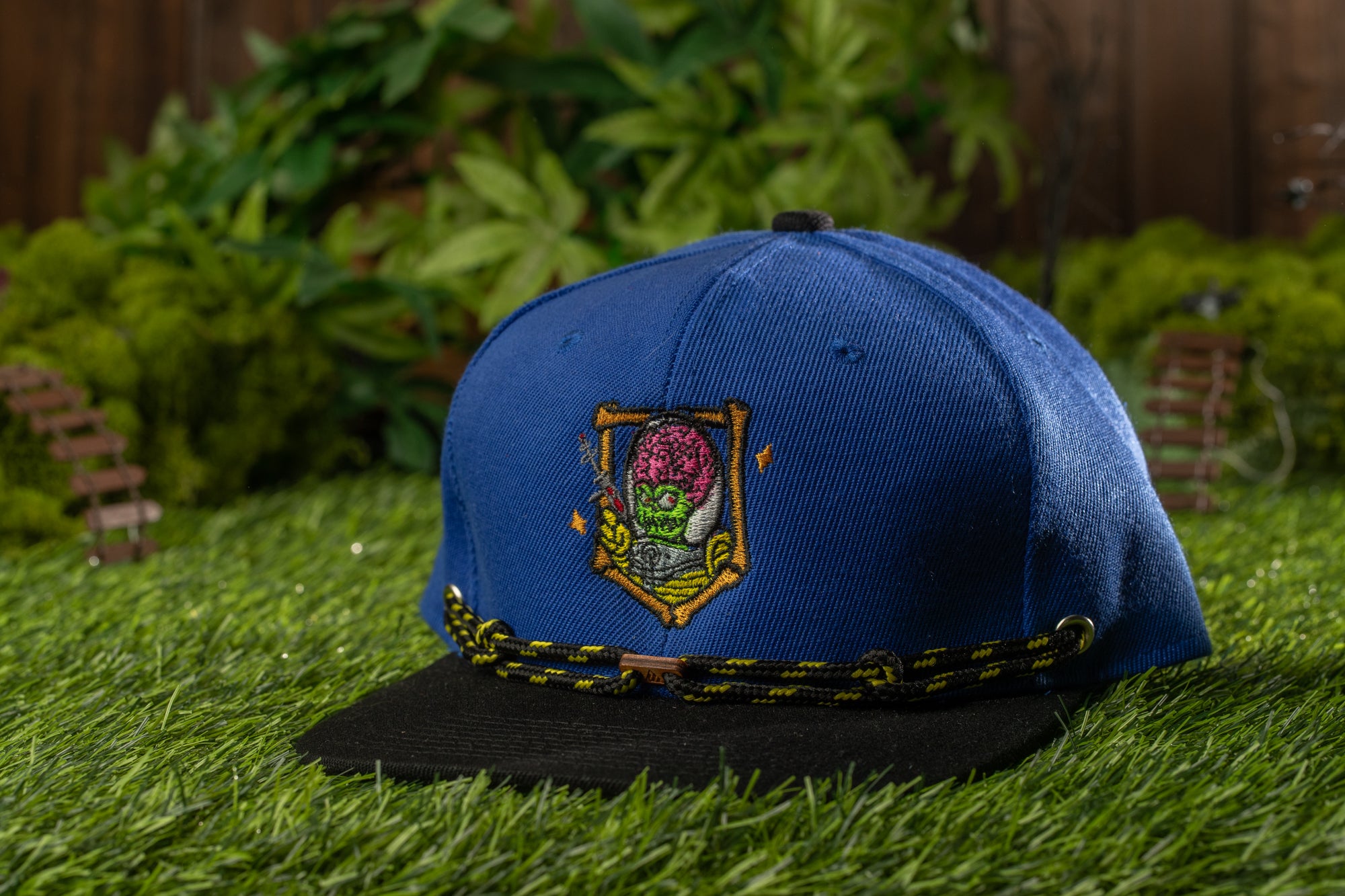 A blue baseball cap with a colorful graphic design sits on a grassy surface, surrounded by a miniature garden scene.