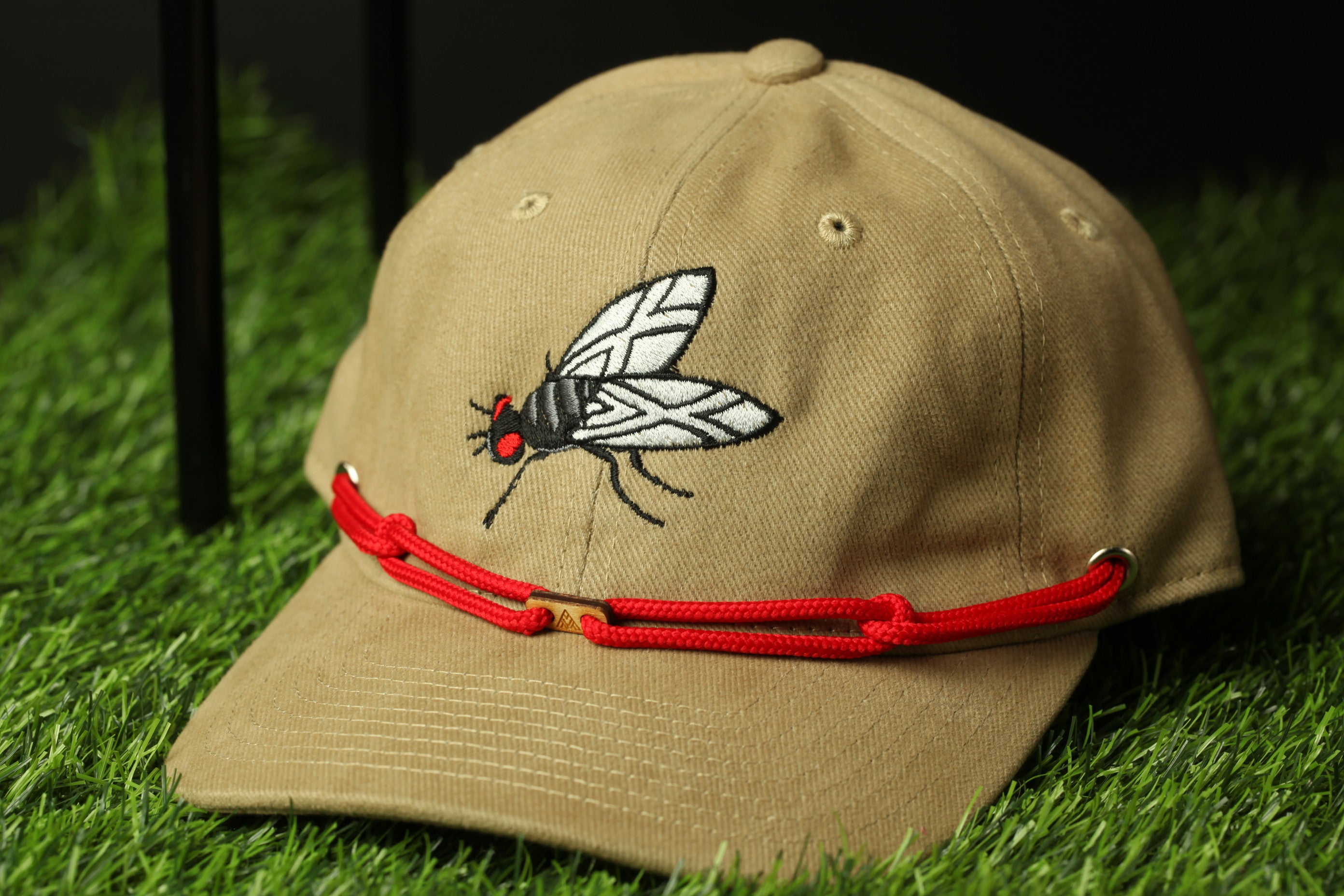 A tan baseball cap with a red string and a black and white fly embroidered on the front, resting on a grassy surface.