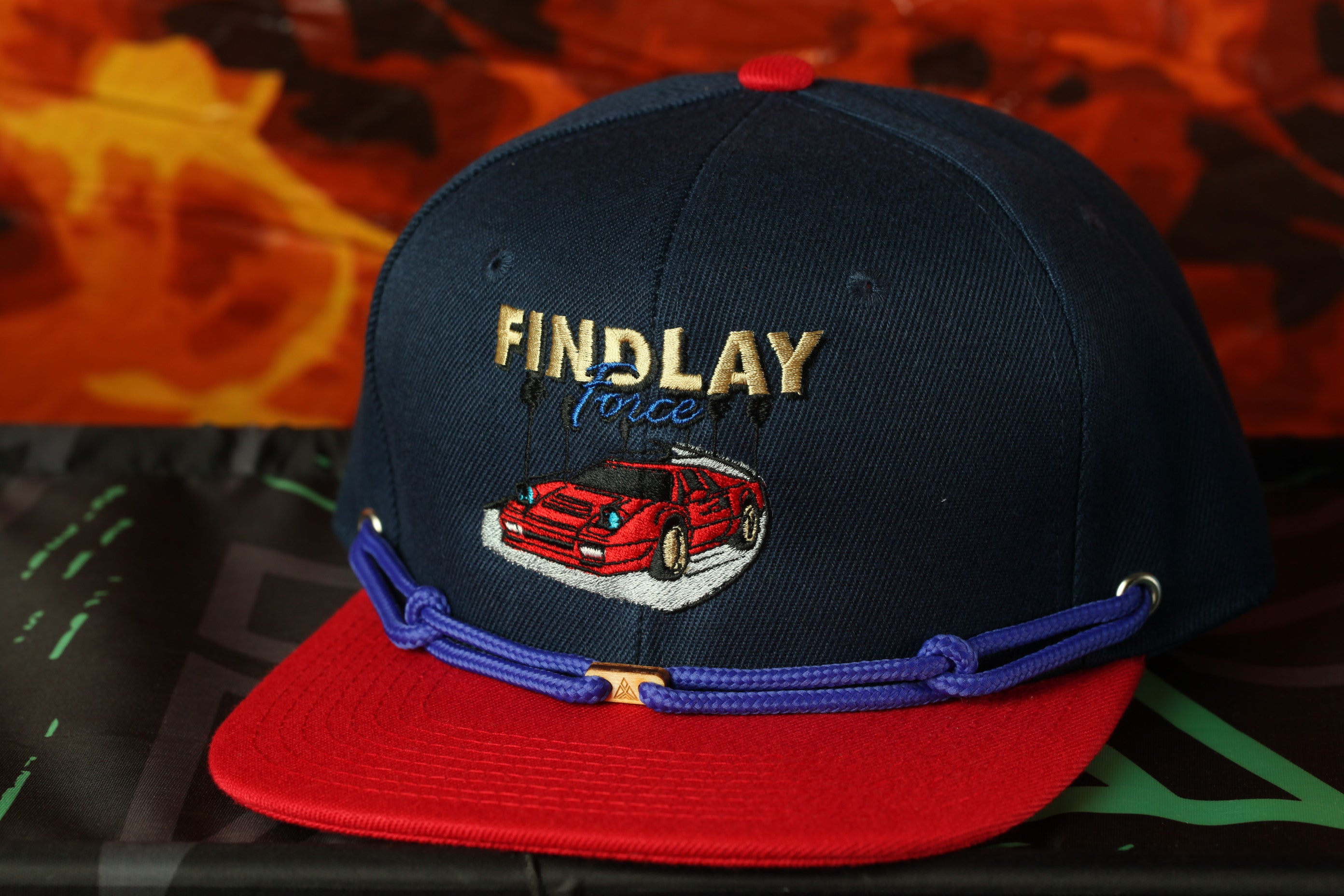 A navy blue baseball cap with a red brim and a red and blue chain strap, featuring an embroidered image of a red sports car.