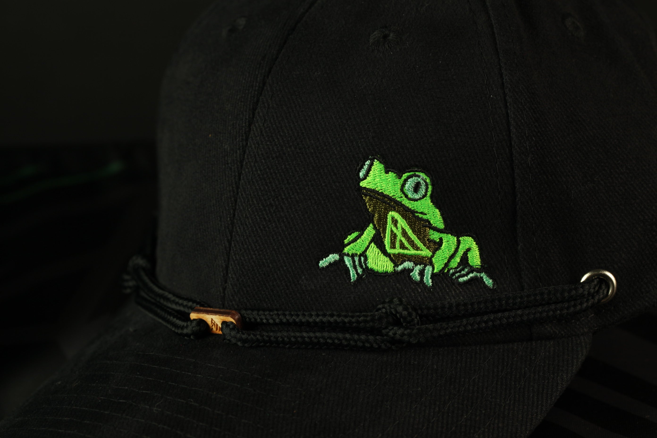 A close-up view of a black baseball cap with a green frog embroidered on the front.