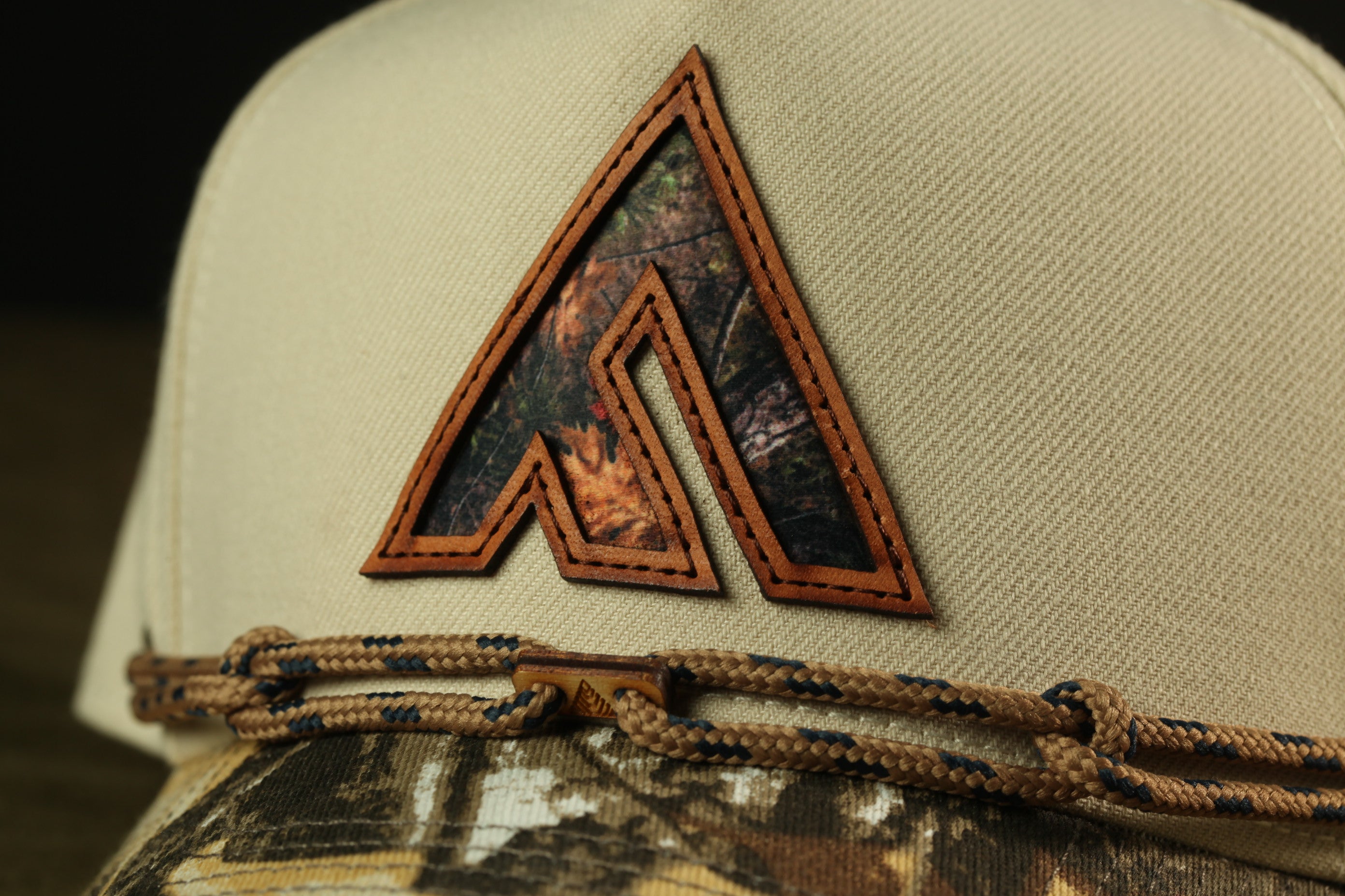 A close-up view of a beige baseball cap with a camouflage pattern and a leather-like logo on the front.