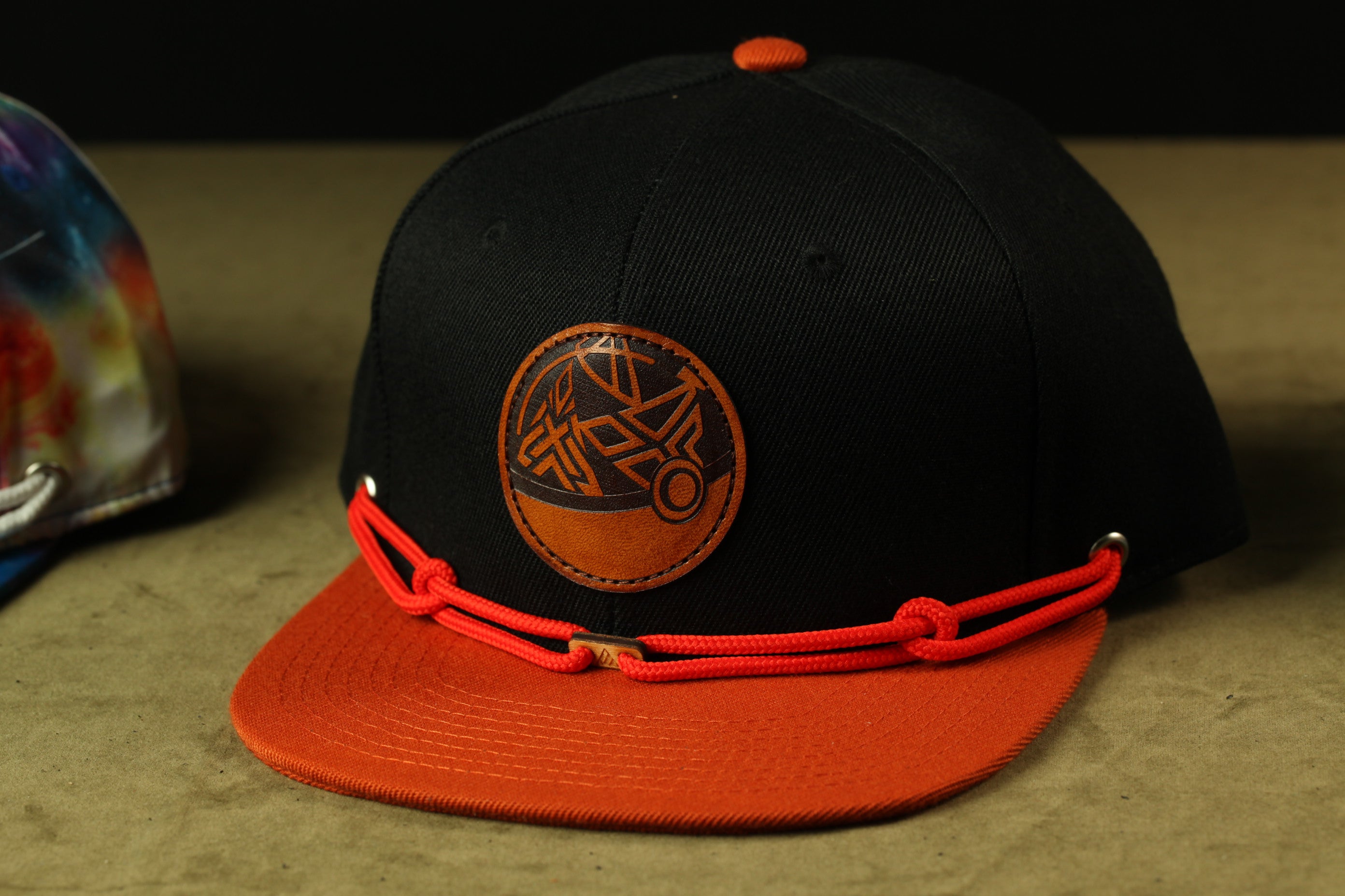 A black baseball cap with an orange brim and a circular logo on the front, sitting on a brown surface.
