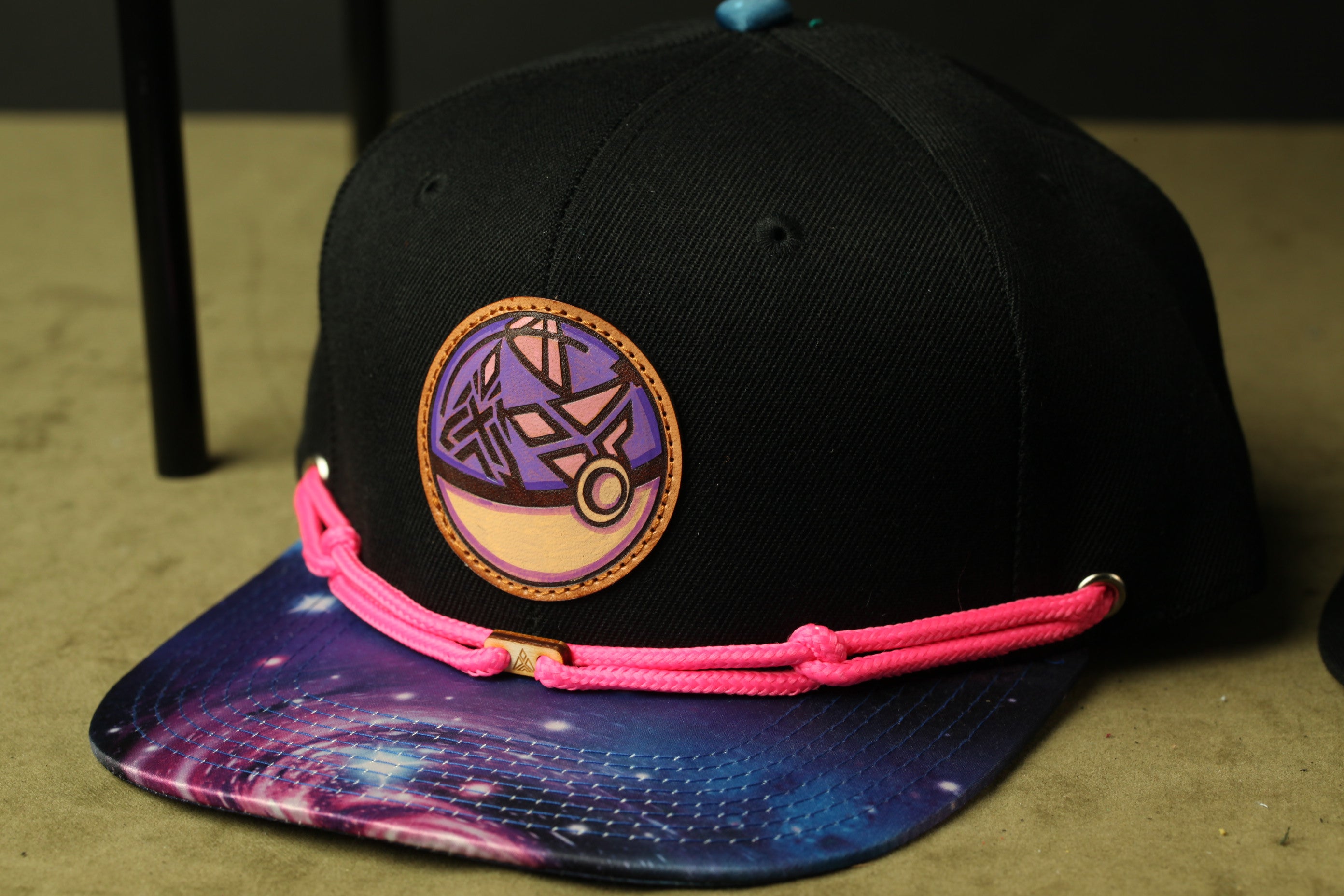 A black baseball cap with a colorful, abstract design on the front, featuring a purple and orange circular motif.