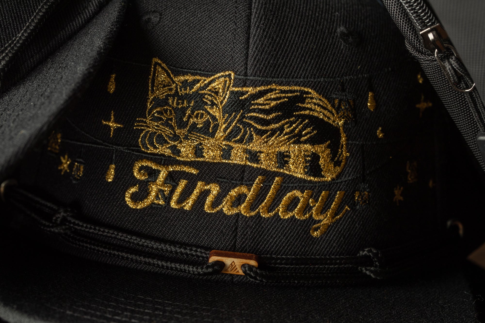 A close-up view of a black baseball cap with gold embroidered text and a cat design.
