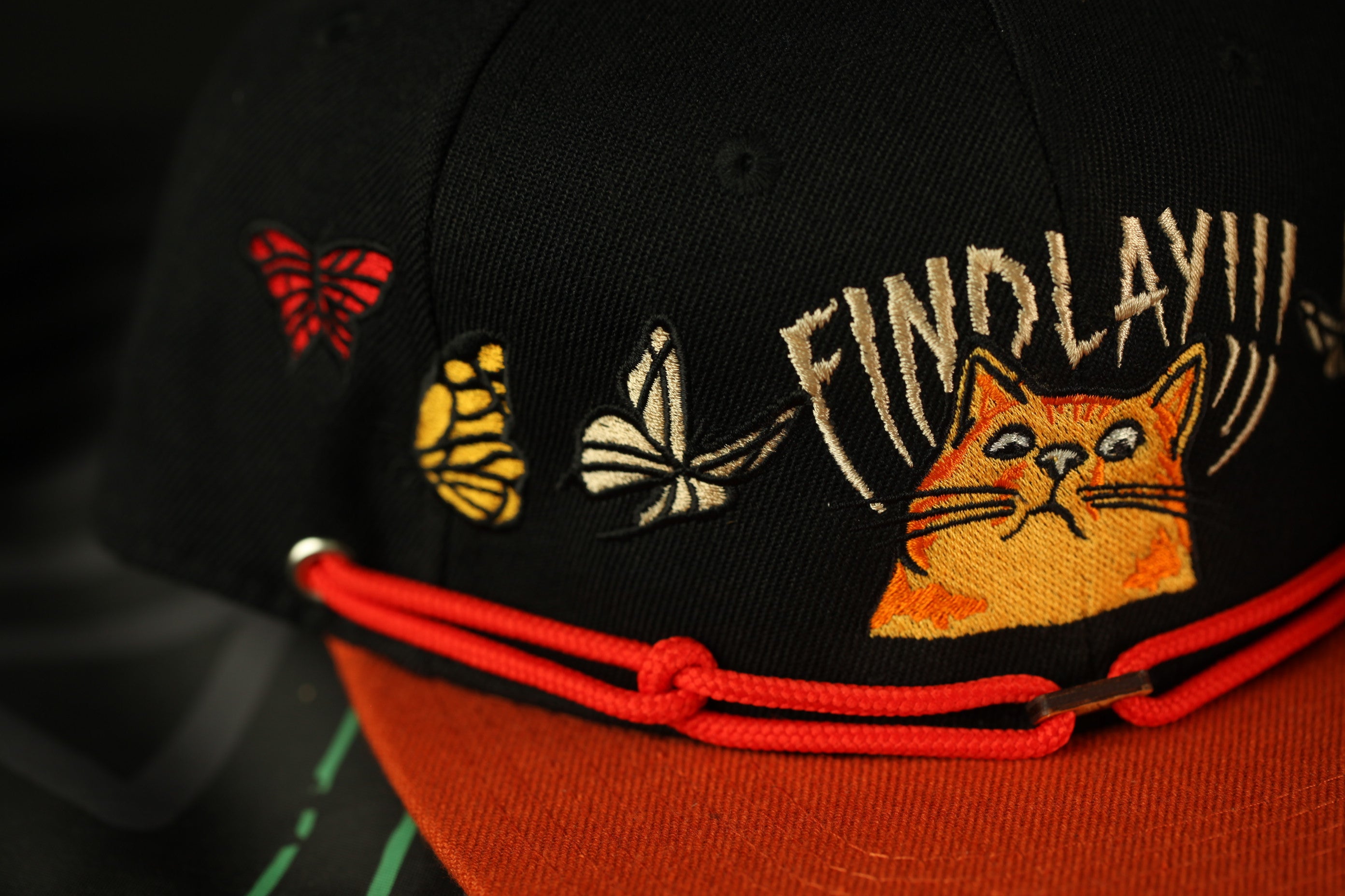 A close-up view of a black baseball cap with various embroidered designs, including a cat, butterflies, and text.
