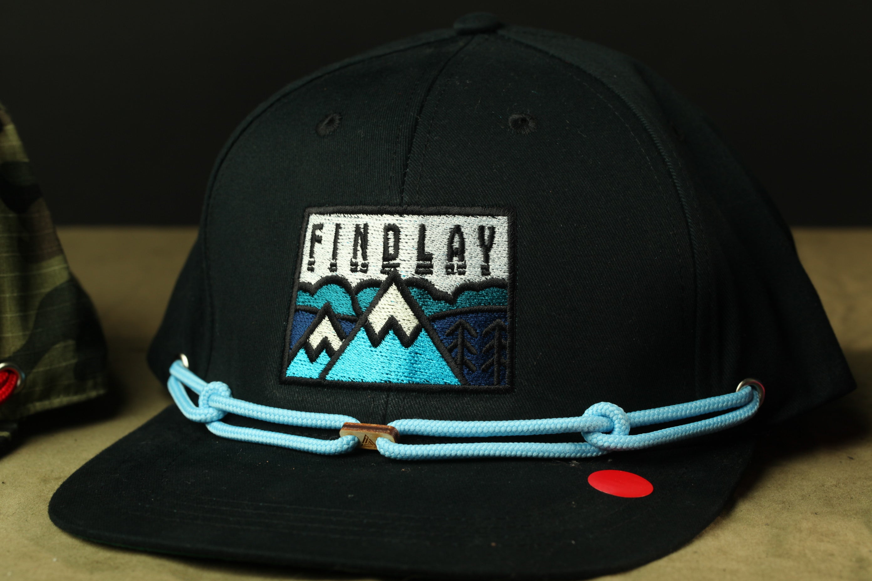A black baseball cap with a blue and white mountain-themed patch and a blue chain strap.