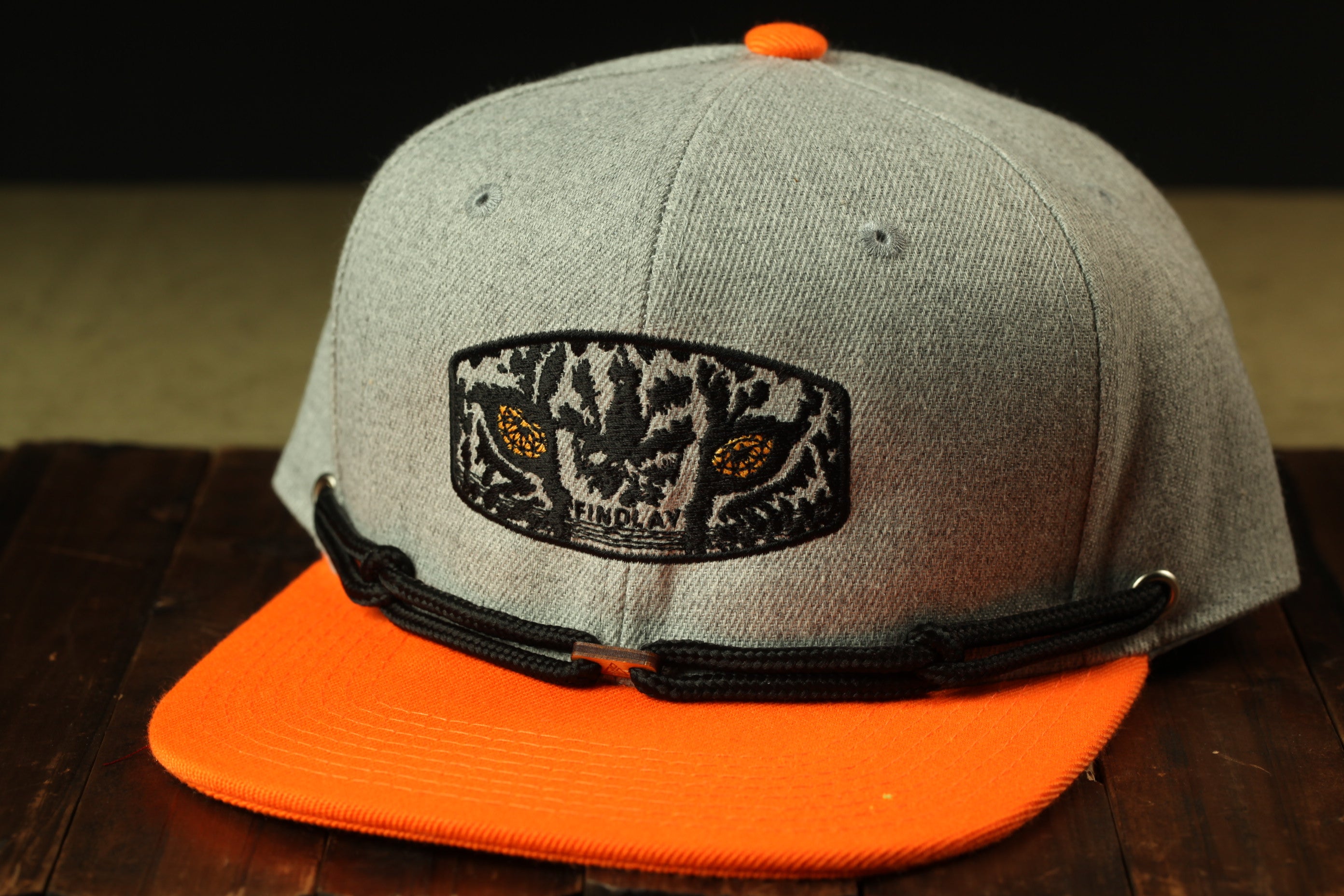A gray baseball cap with an orange brim and a black and white patch on the front, sitting on a wooden surface.