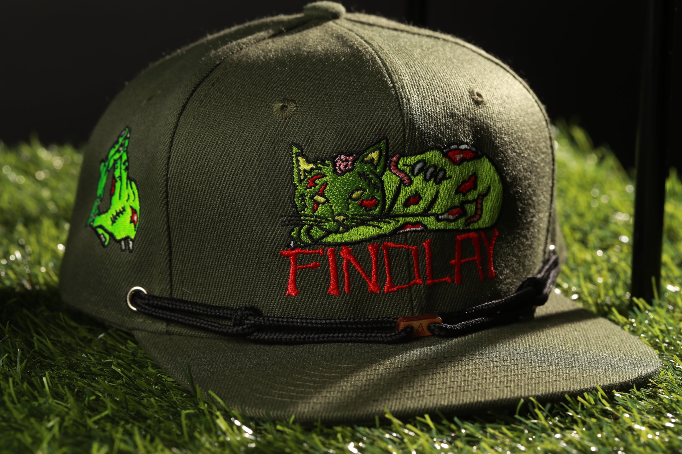 A green baseball cap with a graphic of a green cat and the word "FINDOLAY" printed on it, sitting on a grassy surface.