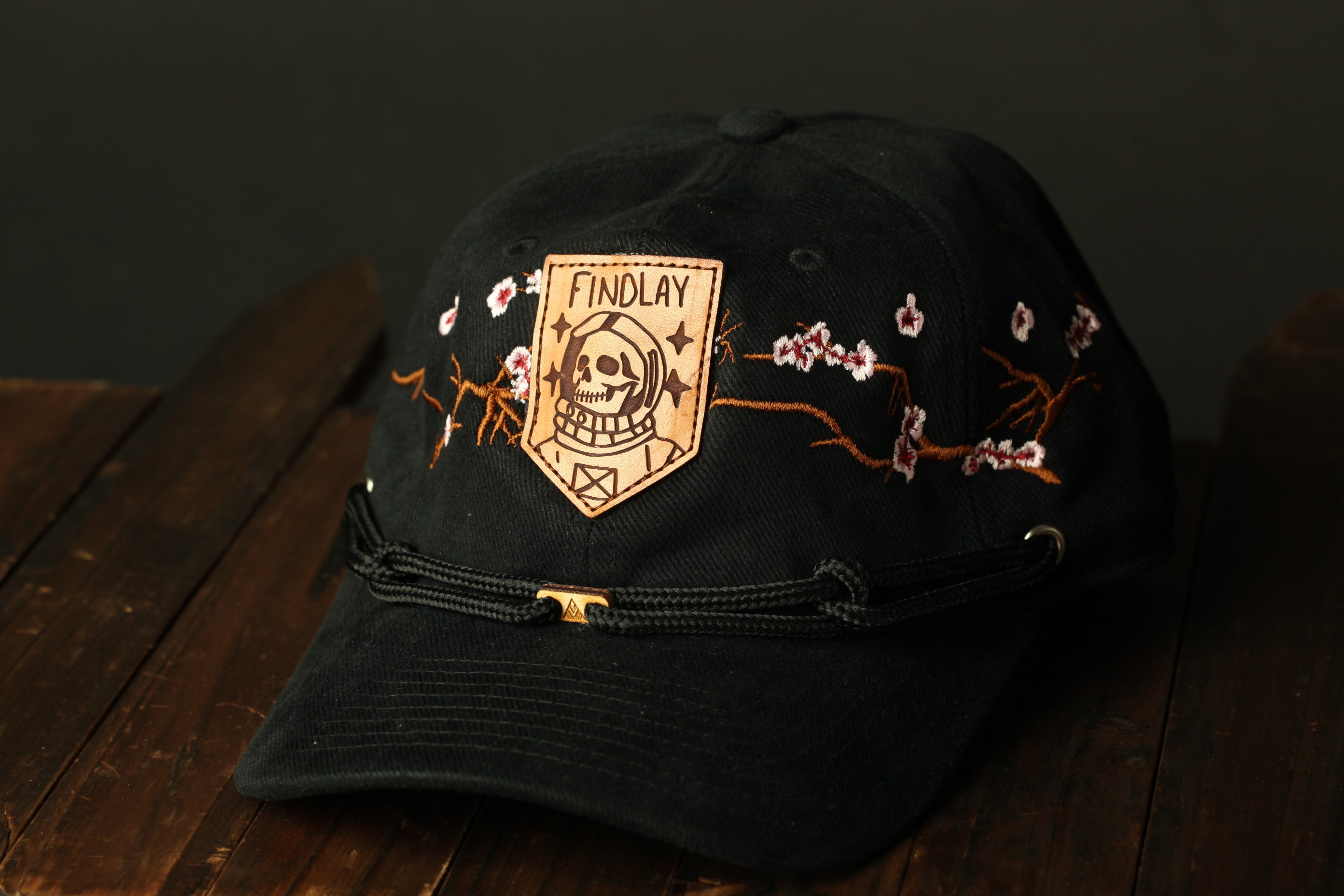 A black baseball cap with a leather patch featuring a skull and floral designs, sitting on a wooden surface.
