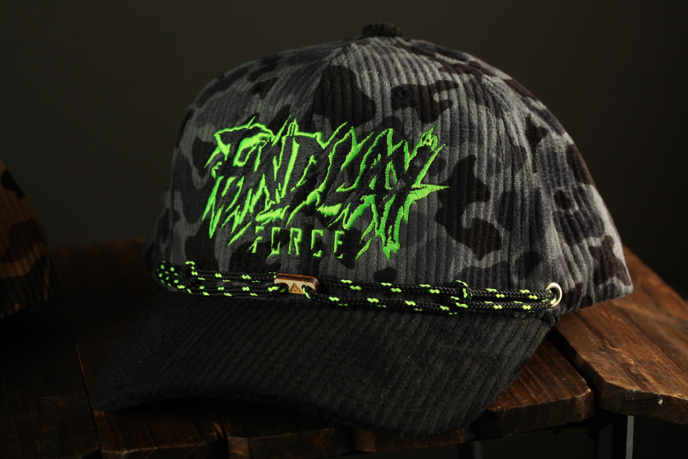 A black and gray camouflage baseball cap with neon green text on it, sitting on a wooden surface.