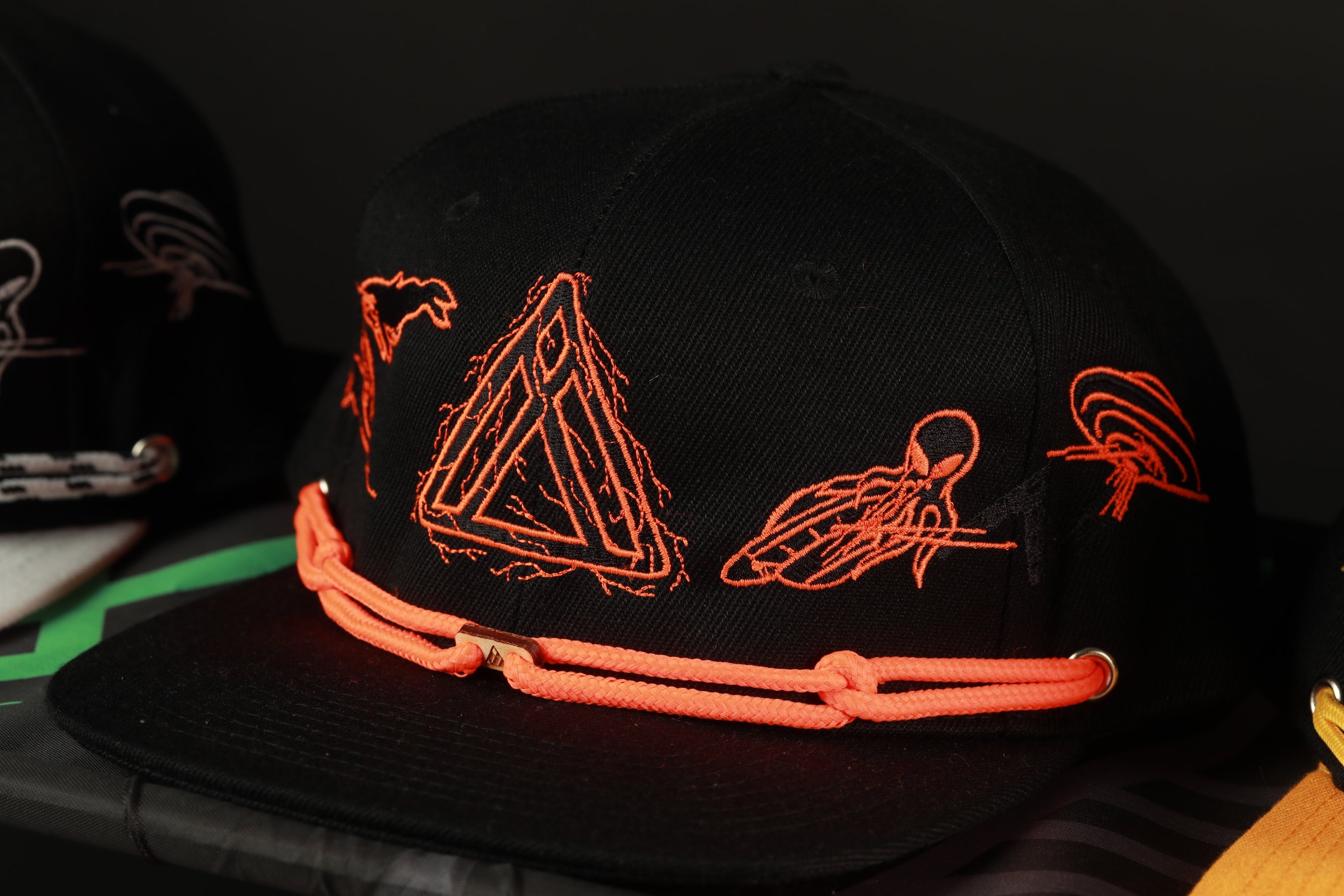 A black baseball cap with orange and black embroidered designs, including a triangle, a fish, and a bird, is displayed on a surface.