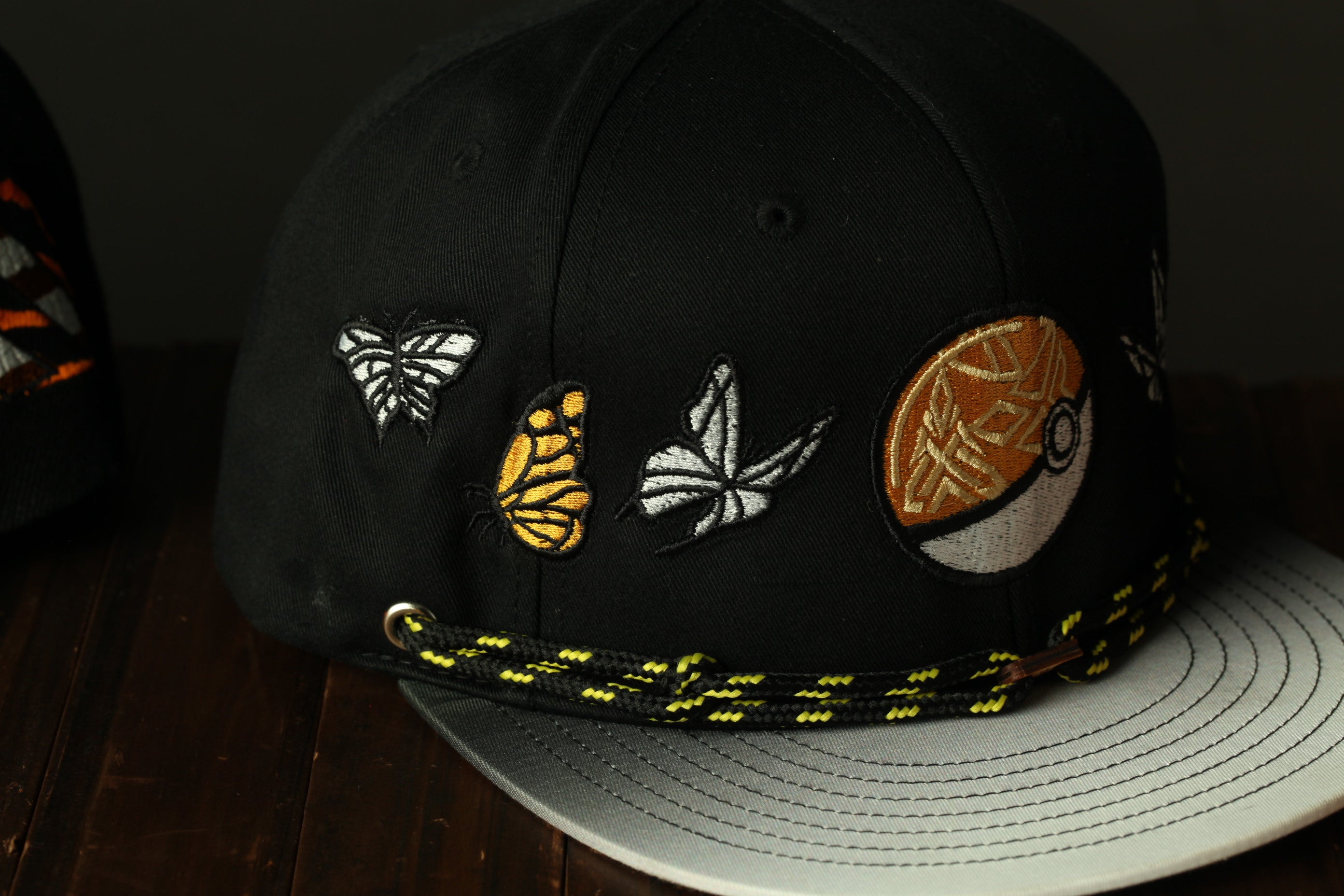A black baseball cap with various embroidered designs, including butterflies and a Pokemon ball, is shown on a wooden surface.