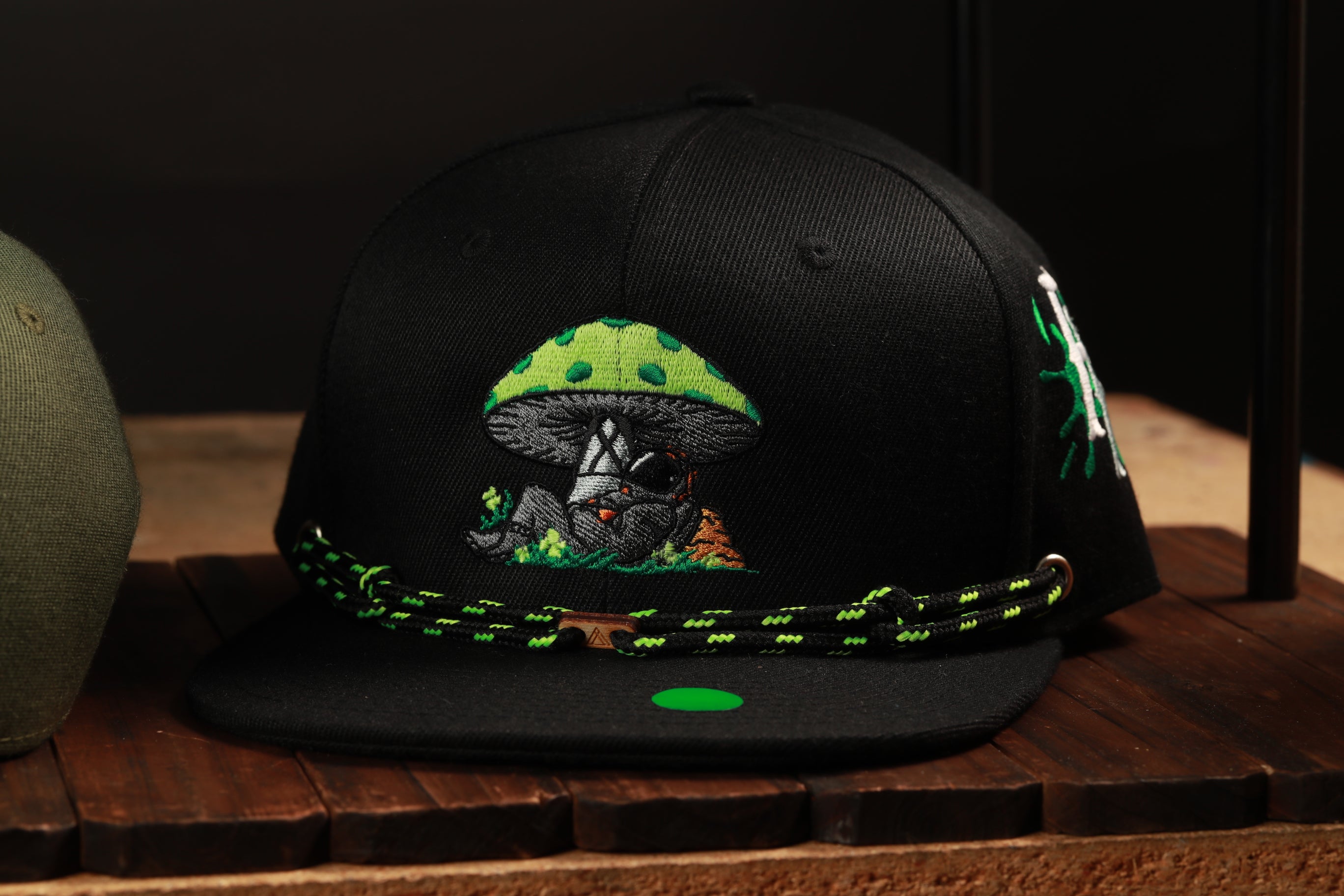A black baseball cap with a green mushroom design and a green string around the brim.