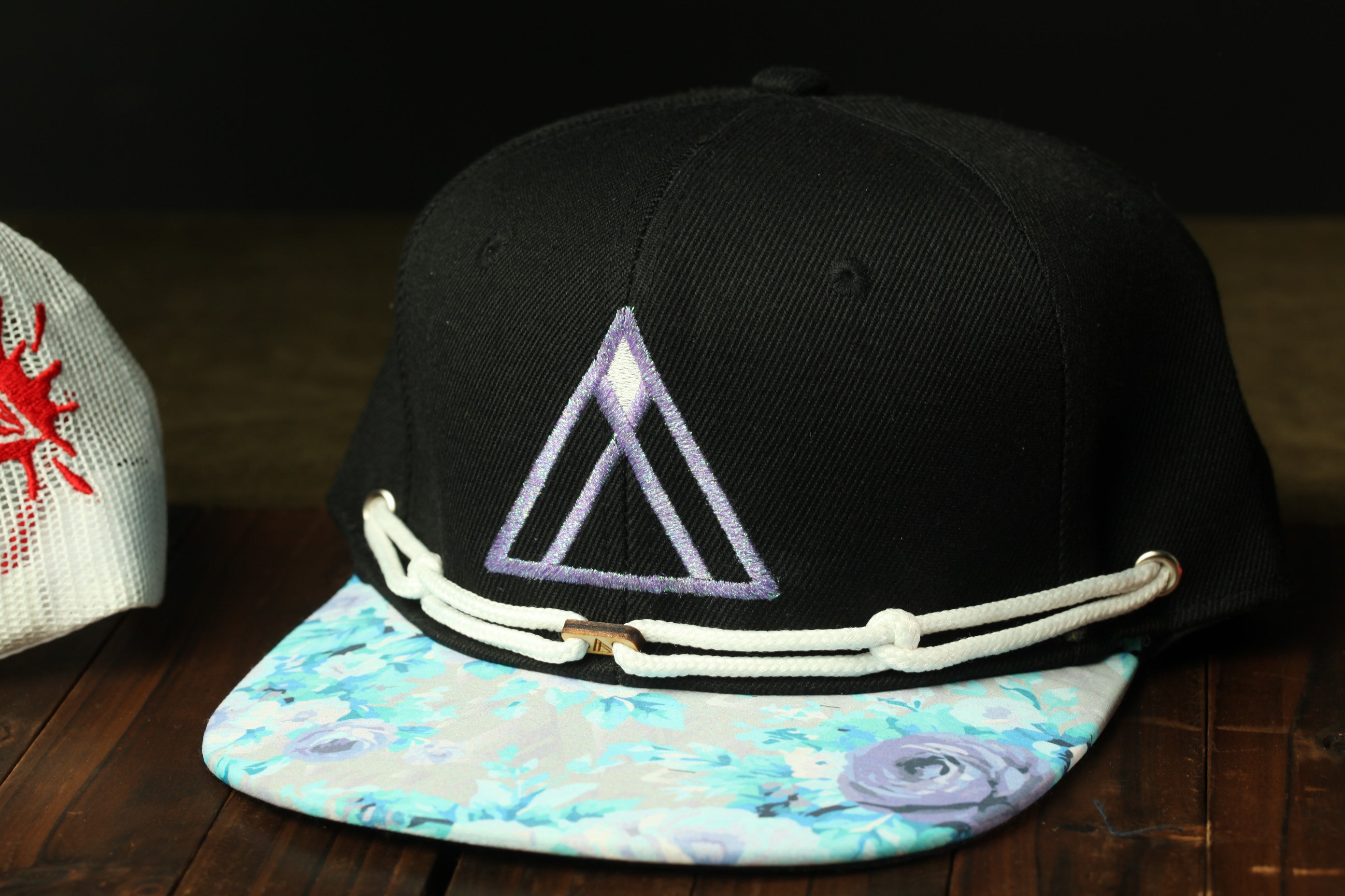 A black baseball cap with a purple triangle design and a floral pattern on the brim, placed on a wooden surface.