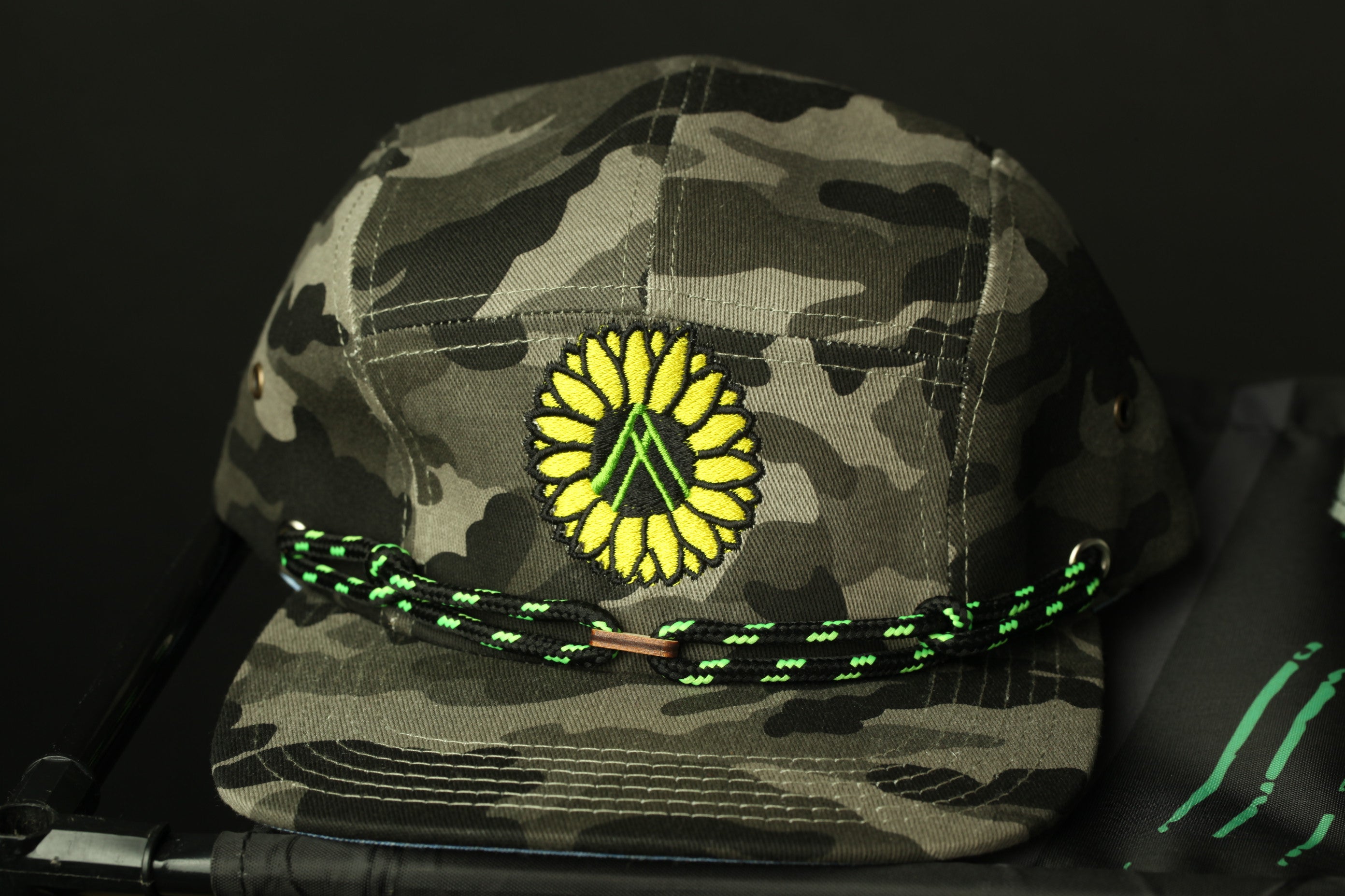 A camouflage baseball cap with a yellow flower embroidered on it, featuring a green string around the brim.