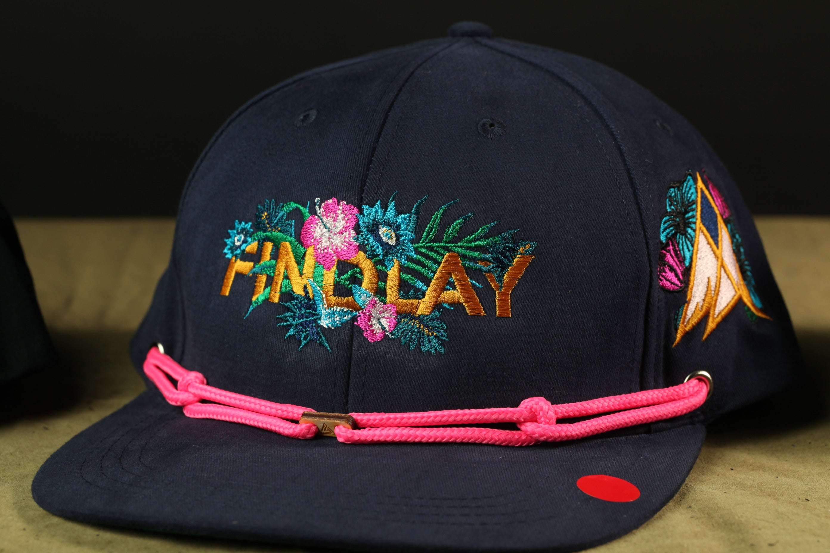 A navy blue baseball cap with a floral embroidered design and a pink braided strap.