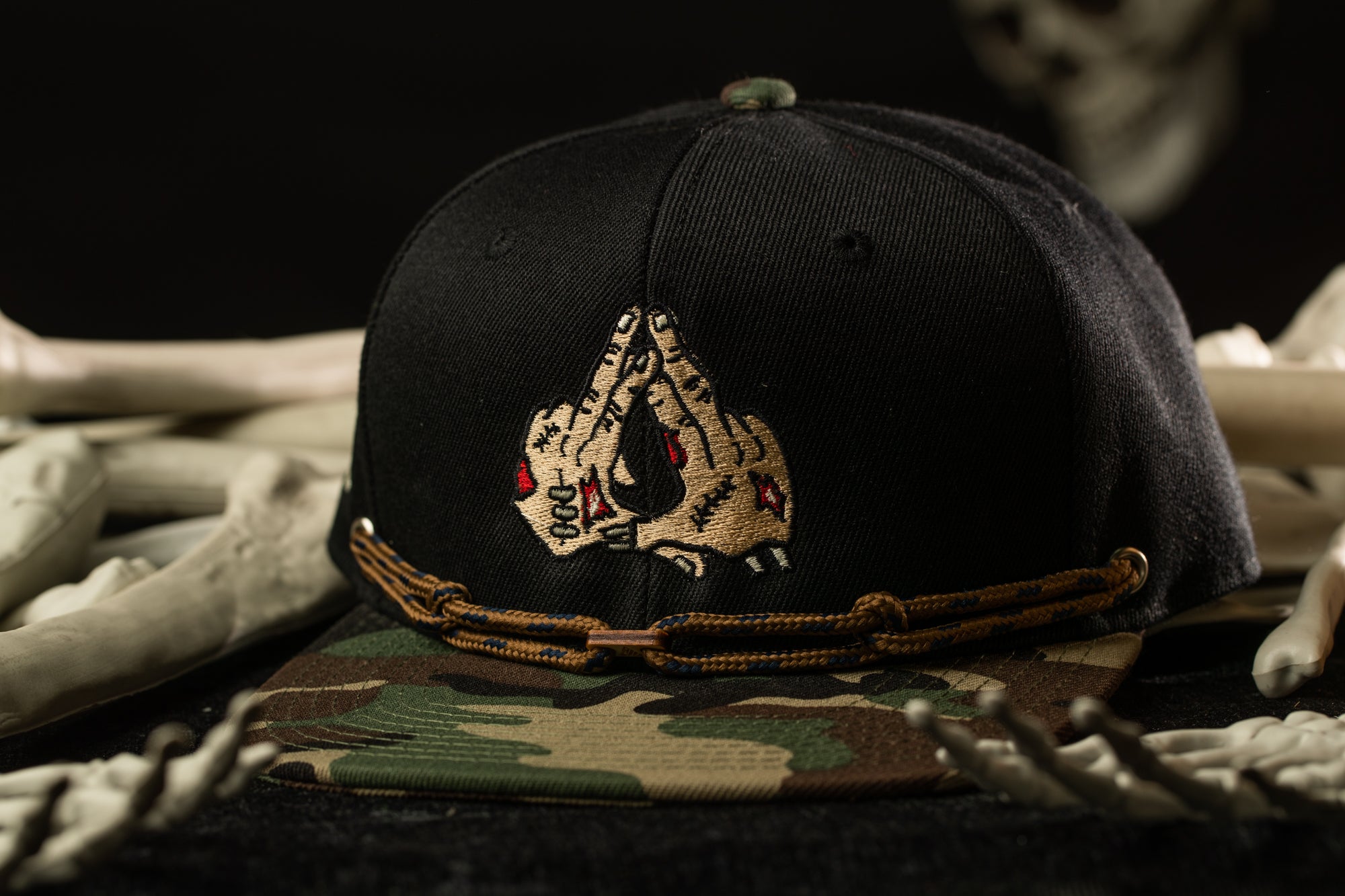 A black baseball cap with a camouflage pattern and a skull design on the front, placed on a surface with skeletal bones.