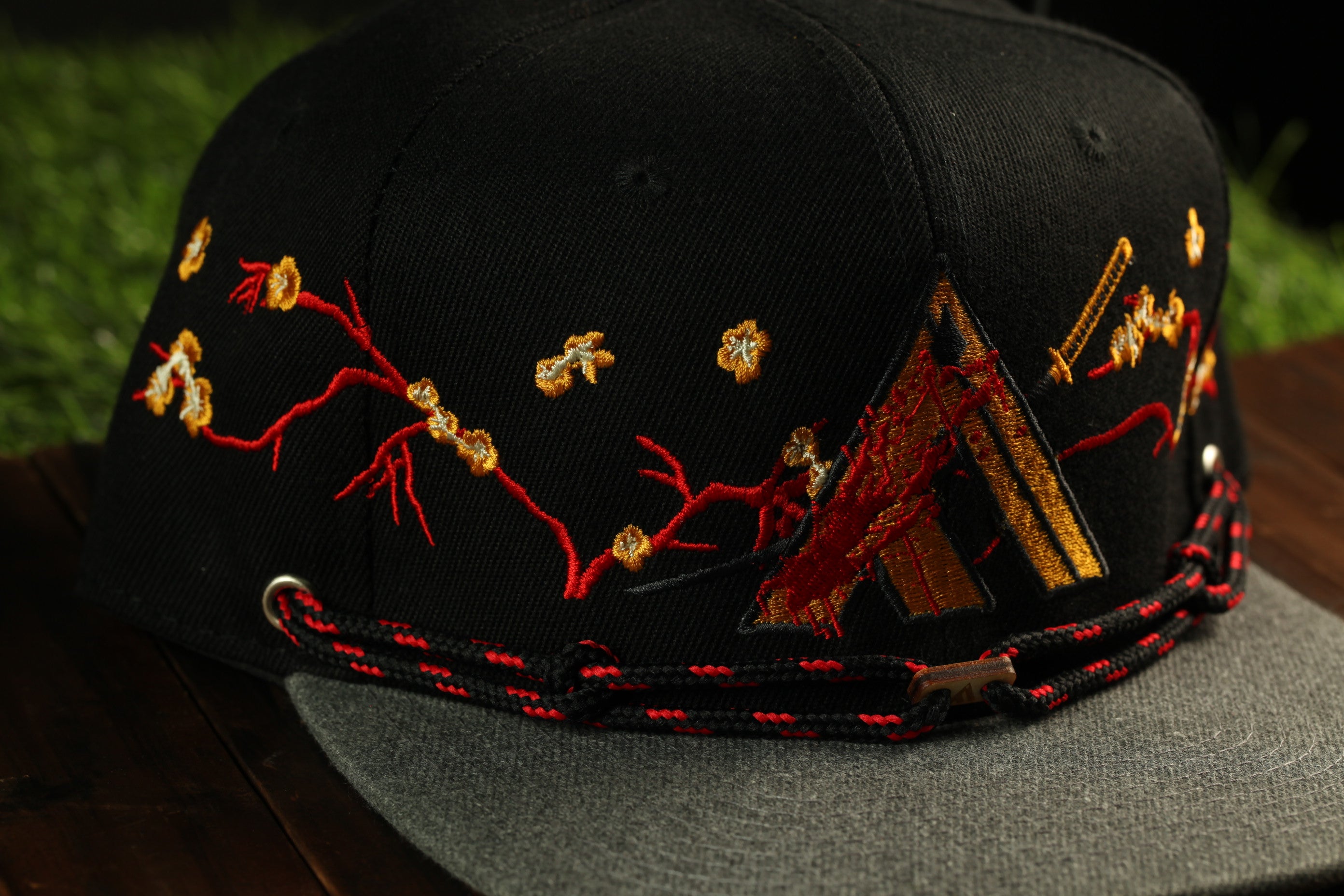 A black baseball cap with red and gold embroidered designs, including a floral pattern and a dragon motif, is shown on a wooden surface.