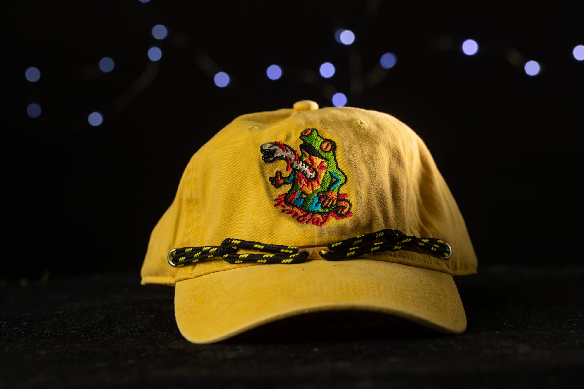 A yellow baseball cap with a colorful frog embroidered on it, set against a dark background with twinkling lights.