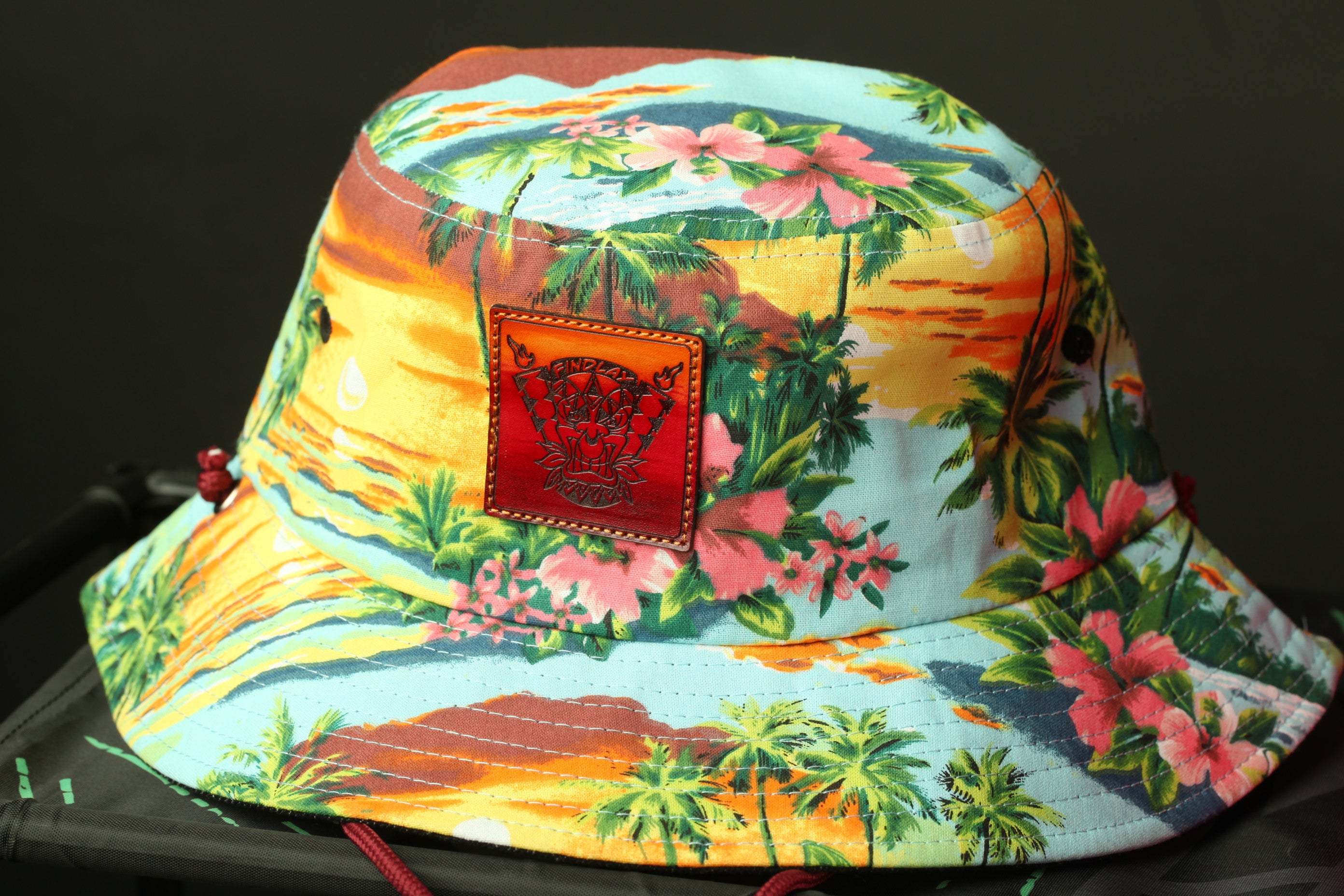 A colorful bucket hat with a tropical print pattern, featuring palm trees, flowers, and a sunset scene.