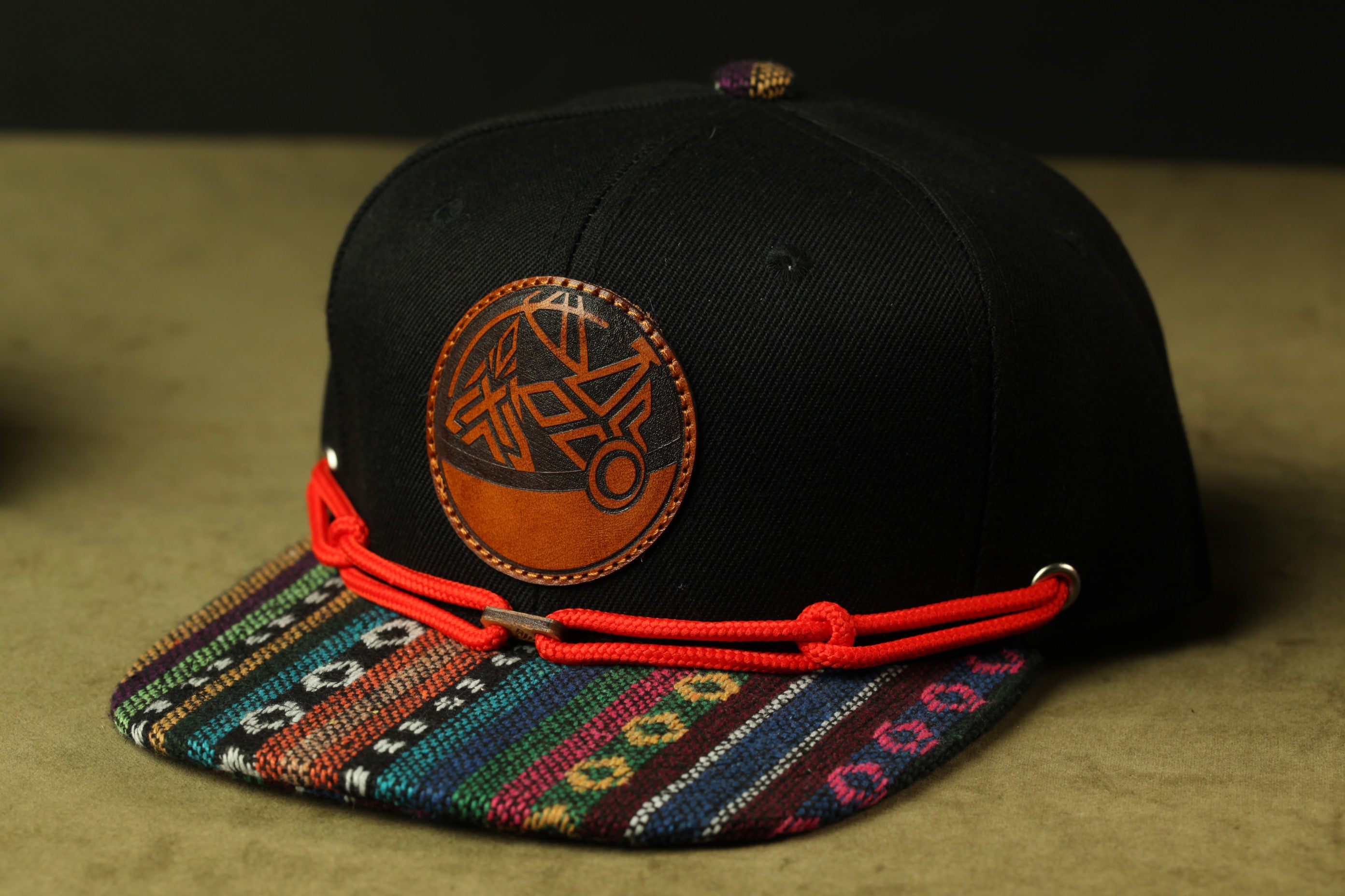 A black baseball cap with a colorful, patterned fabric trim and a circular leather patch on the front.
