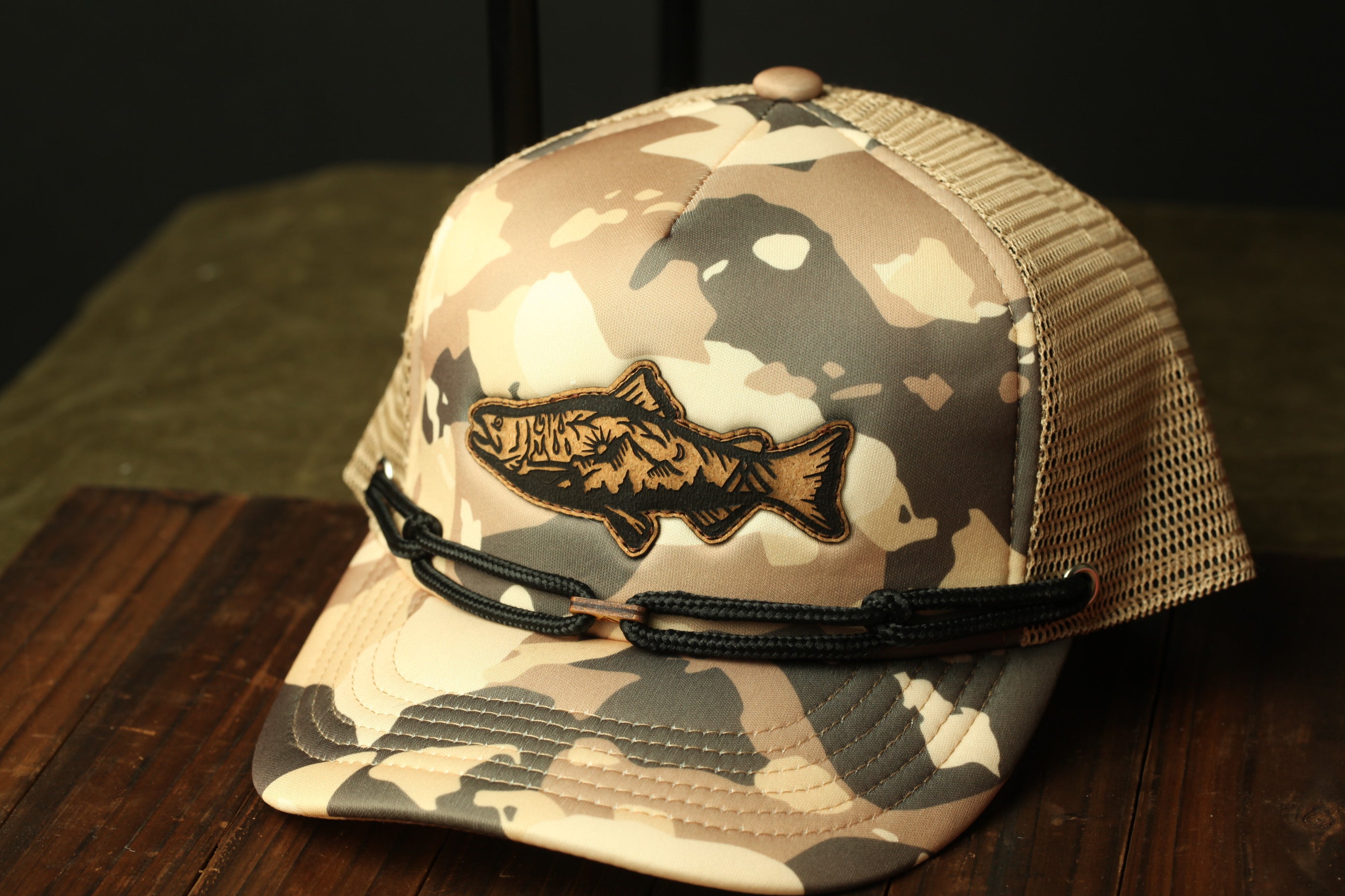 A camouflage baseball cap with a fish embroidered on it, resting on a wooden surface.