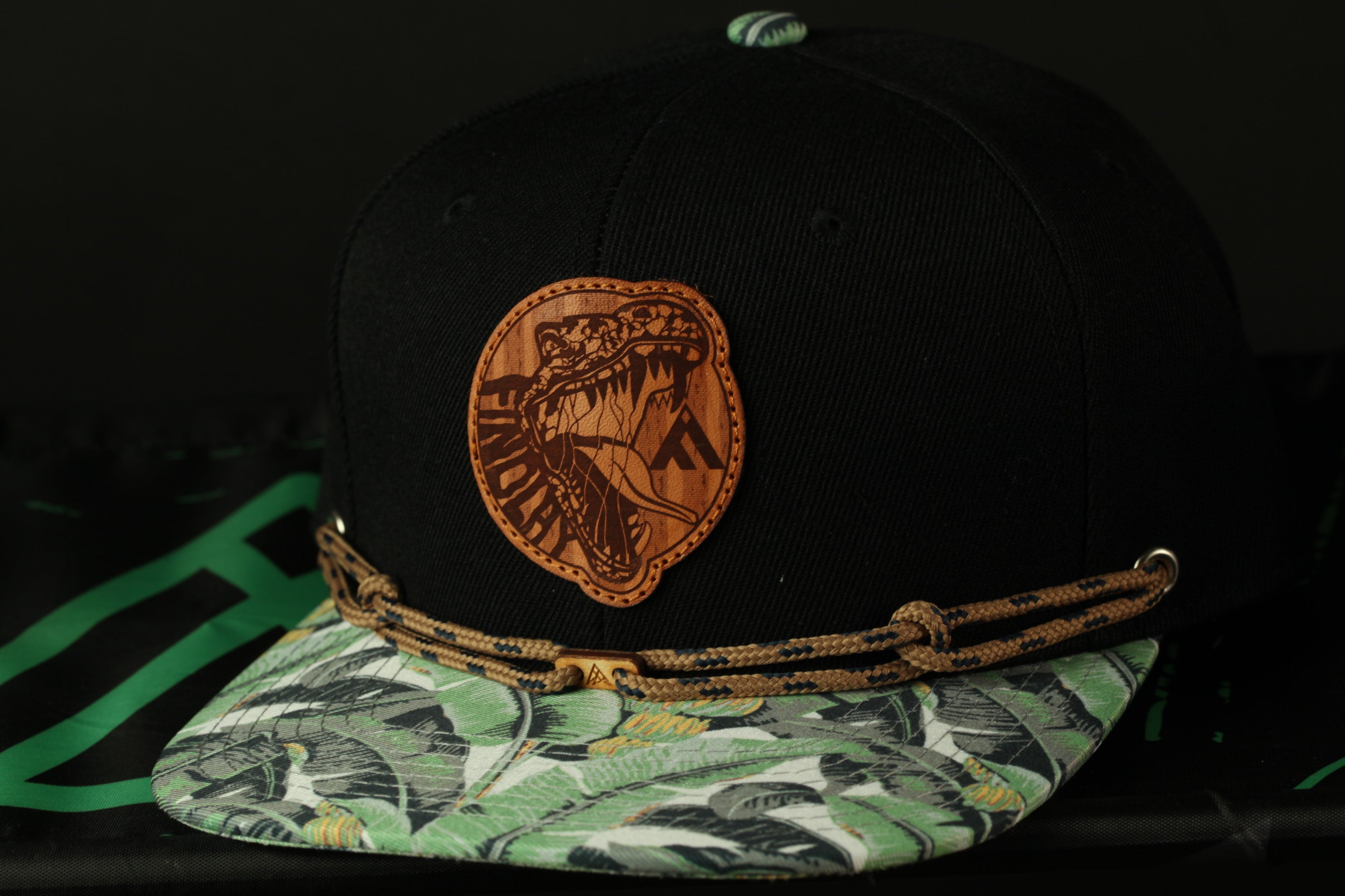 A black baseball cap with a brown leather patch featuring a stylized design, including a snake or alligator, on the front.