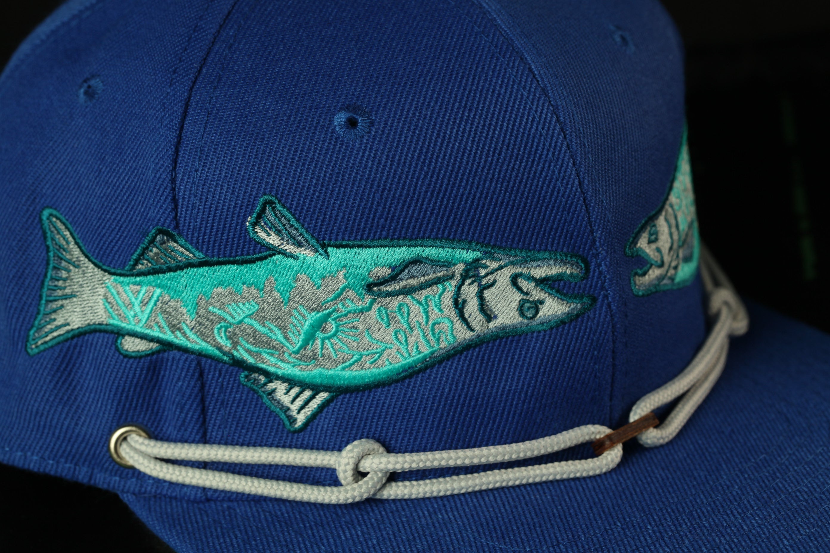 A close-up view of a blue baseball cap with a fish embroidered on it, featuring a detailed design and a white rope strap.