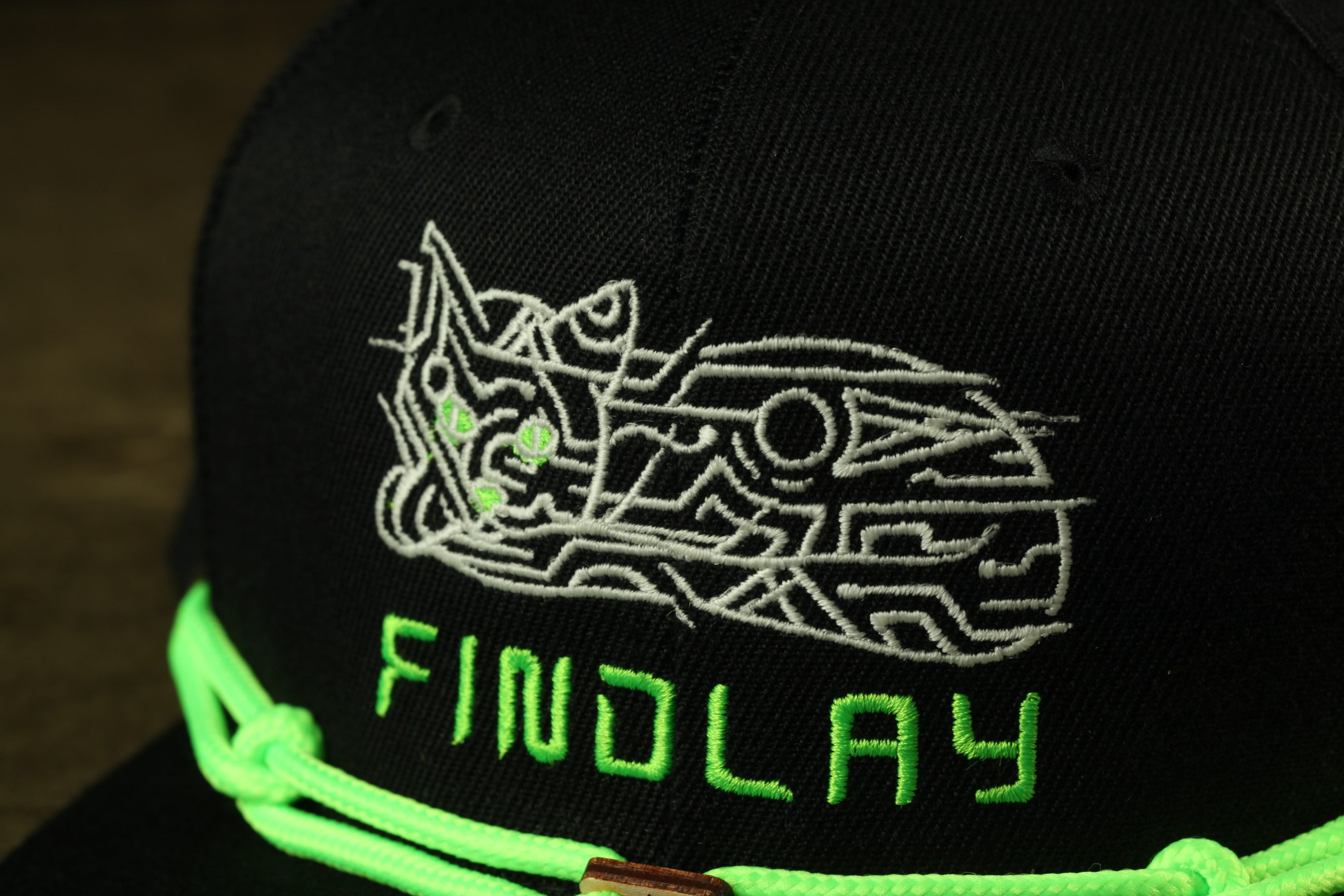 A close-up view of a black baseball cap with a white embroidered design and the word "FINDLAY" written in green on the front.
