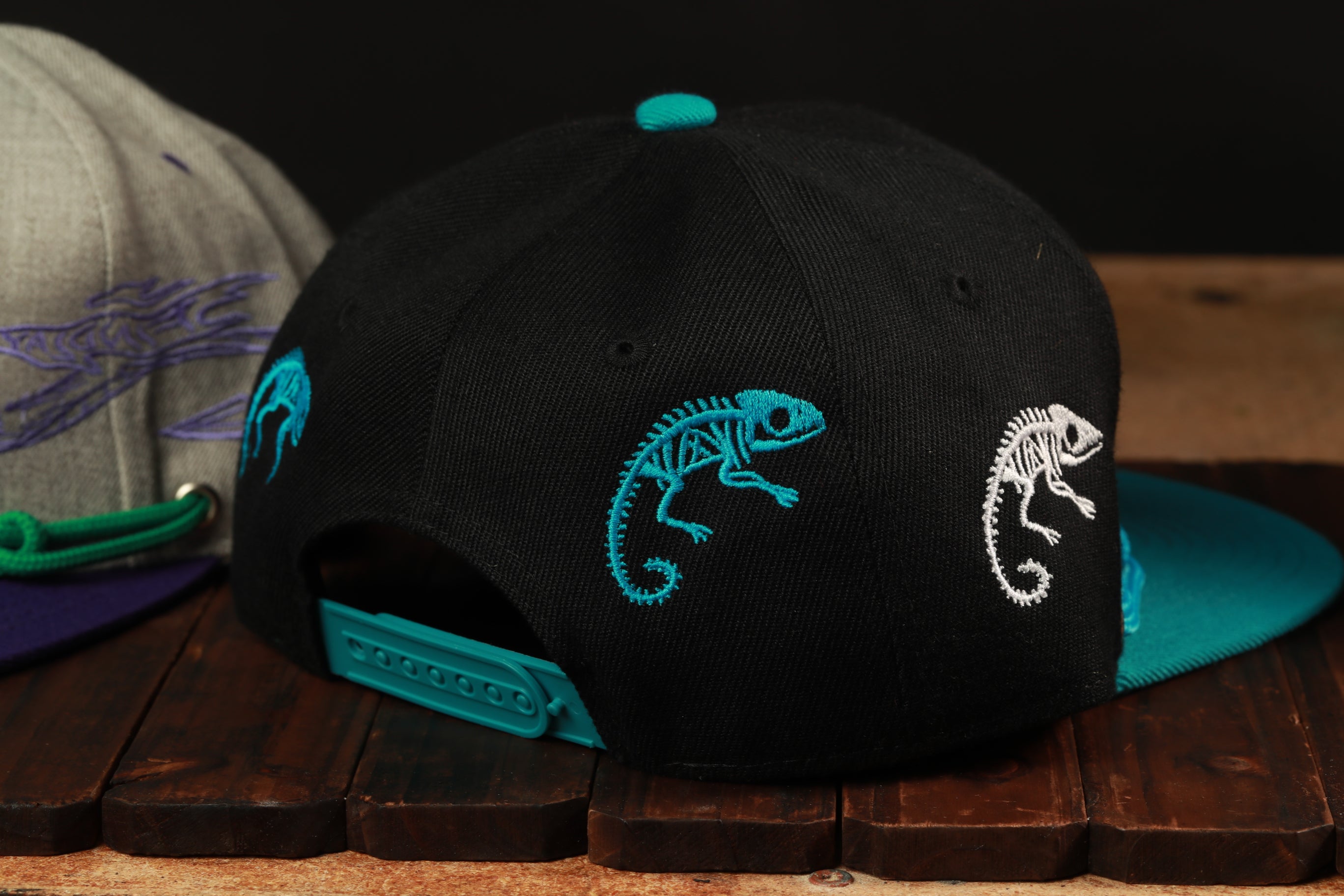 a black baseball cap with blue and teal embroidered designs, including a chameleon and other animal-like patterns, resting on a wooden surface.
