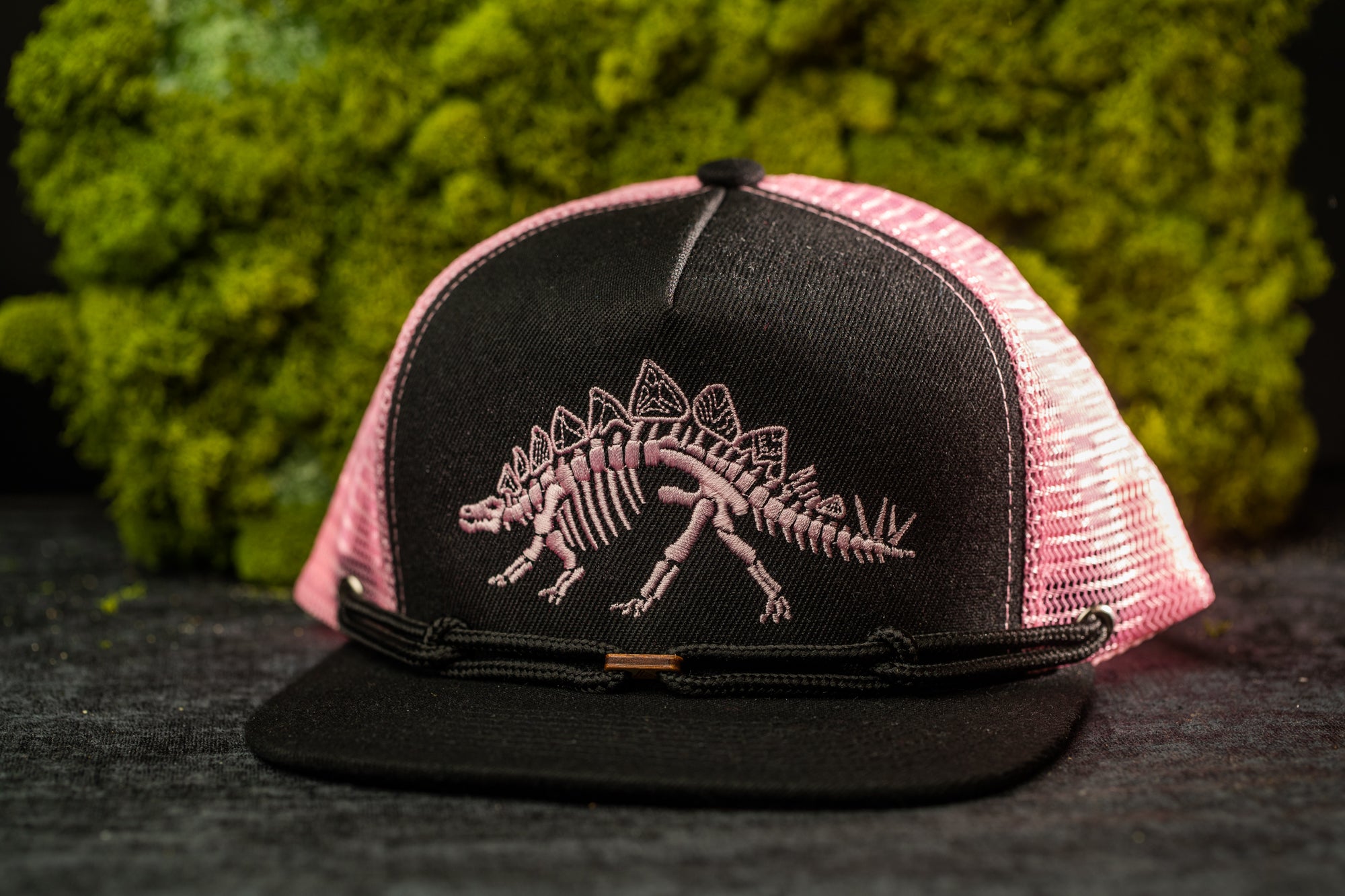 A black baseball cap with a pink dinosaur embroidered on it, placed on a dark surface against a background of green moss.
