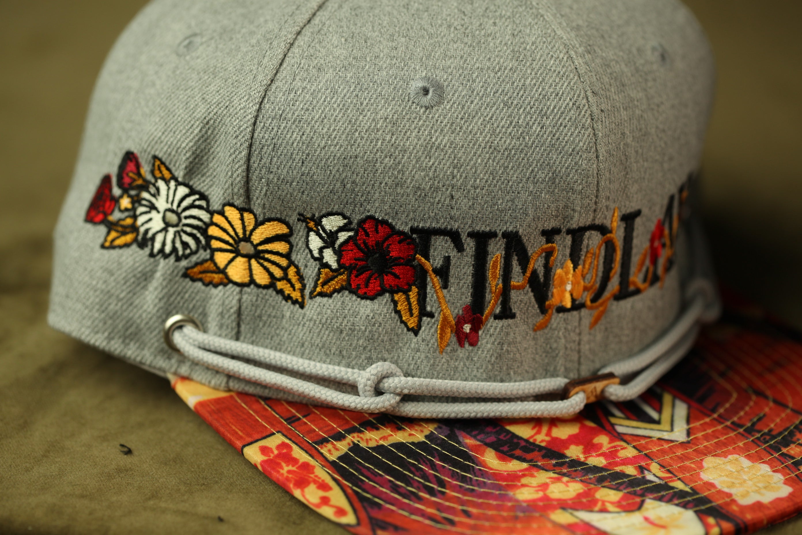 A close-up image of a gray baseball cap with colorful floral embroidery and the word "FIND" printed on it.